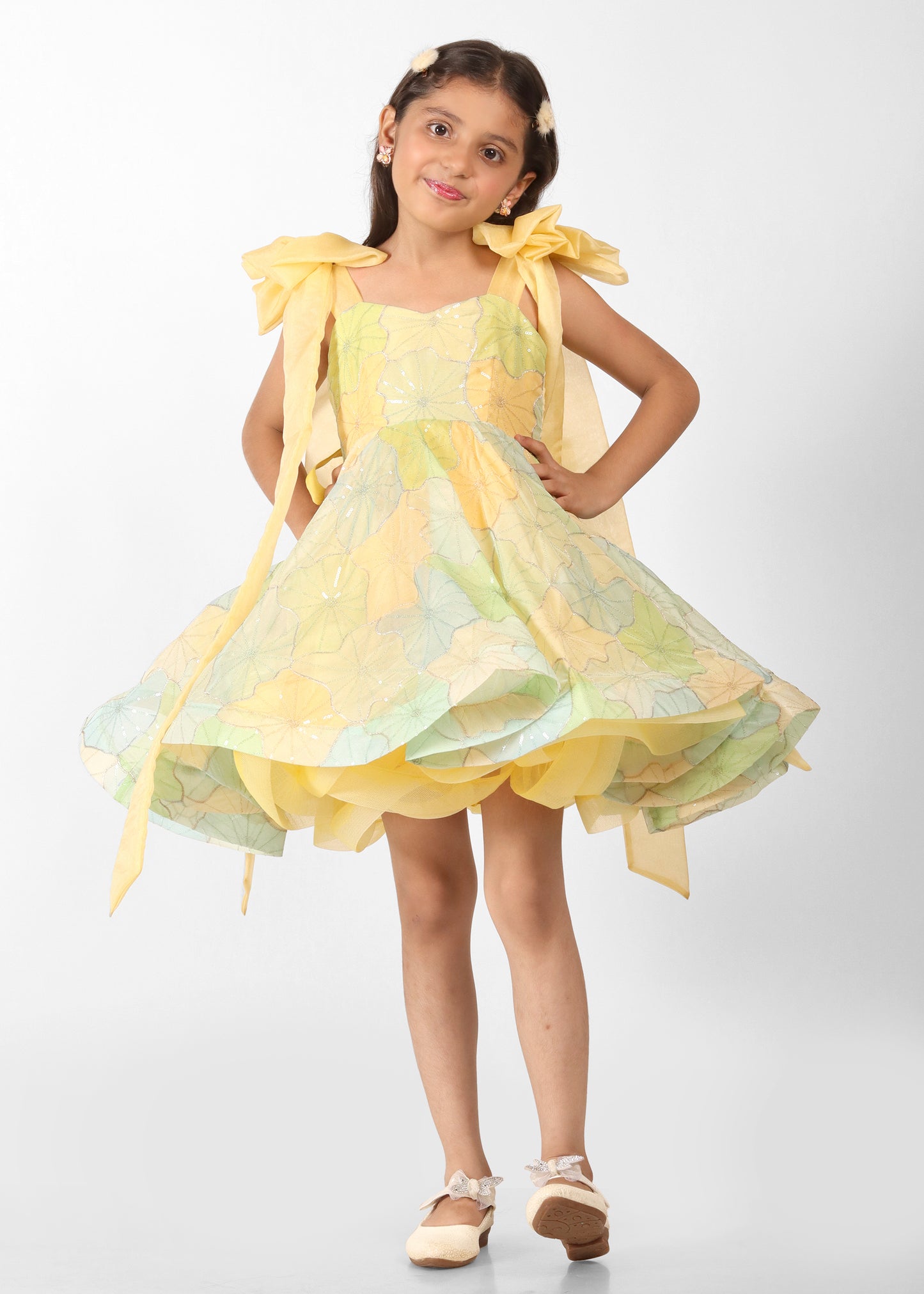 "Full view of a young girl in a pastel yellow floral dress with large bow straps and flared hem, showcasing a fun and playful look."