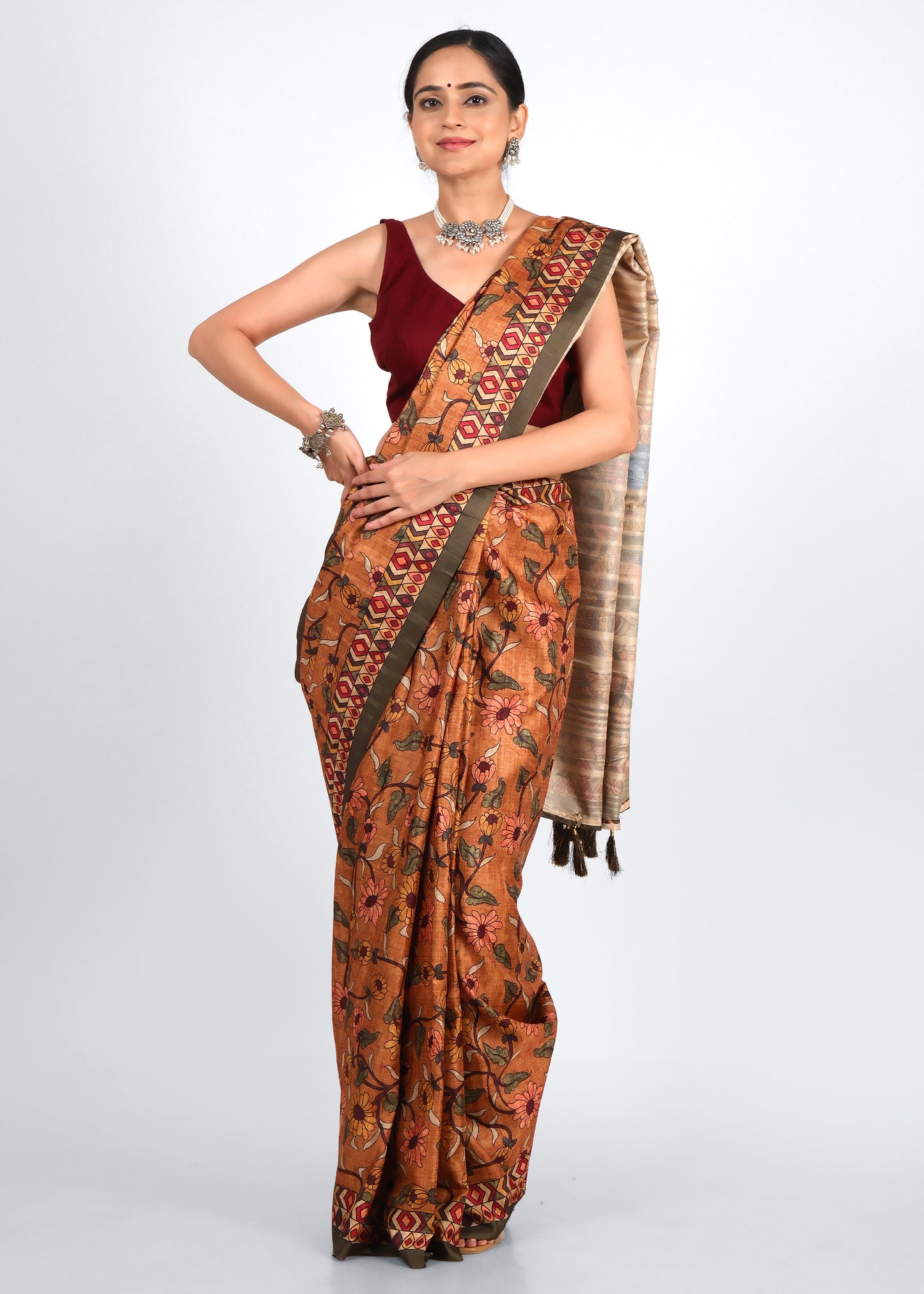 A woman wearing a traditional handloom saree with earthy brown and orange tones, featuring intricate floral patterns and a bold geometric border. The saree is paired with a maroon blouse and silver ethnic jewelry