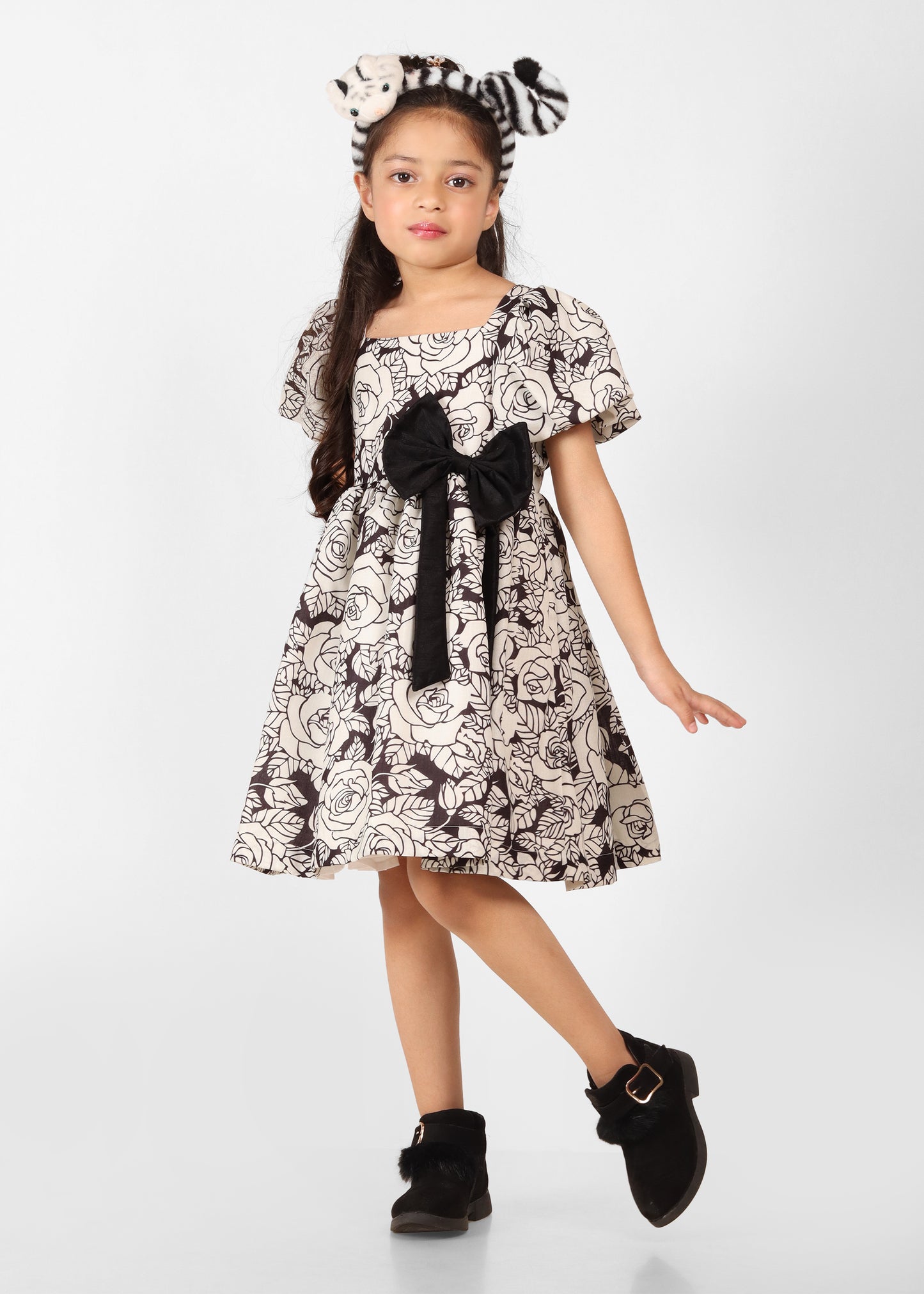 A young girl posing confidently in a black and white floral dress with puffed sleeves and a large black bow at the waist, paired with black ankle boots and a playful headband, showcasing kids' fashion