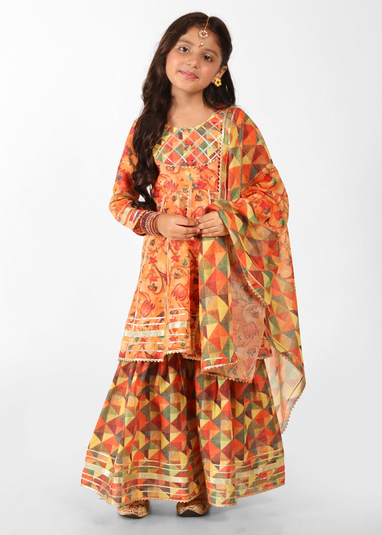 A young girl dressed in a vibrant orange floral printed kurta with a geometric patterned palazzo and matching dupatta. The outfit features intricate embroidery and colorful designs, perfect for traditional Indian festivals and celebrations.