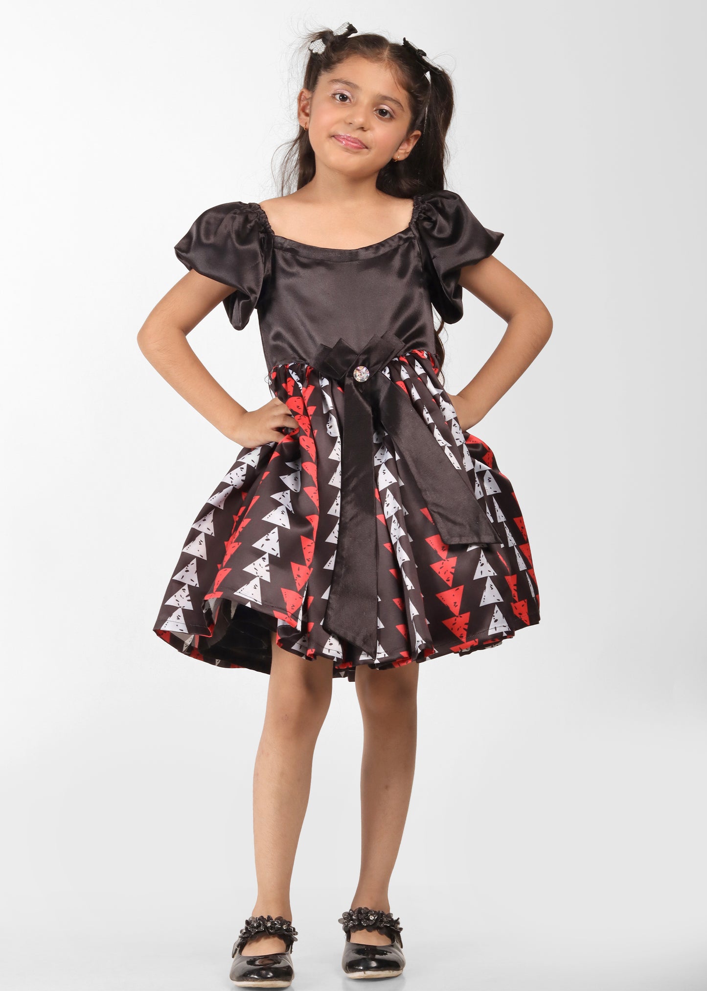 Little girl in a black satin dress with red and white geometric patterns, posing with her hands on her hips