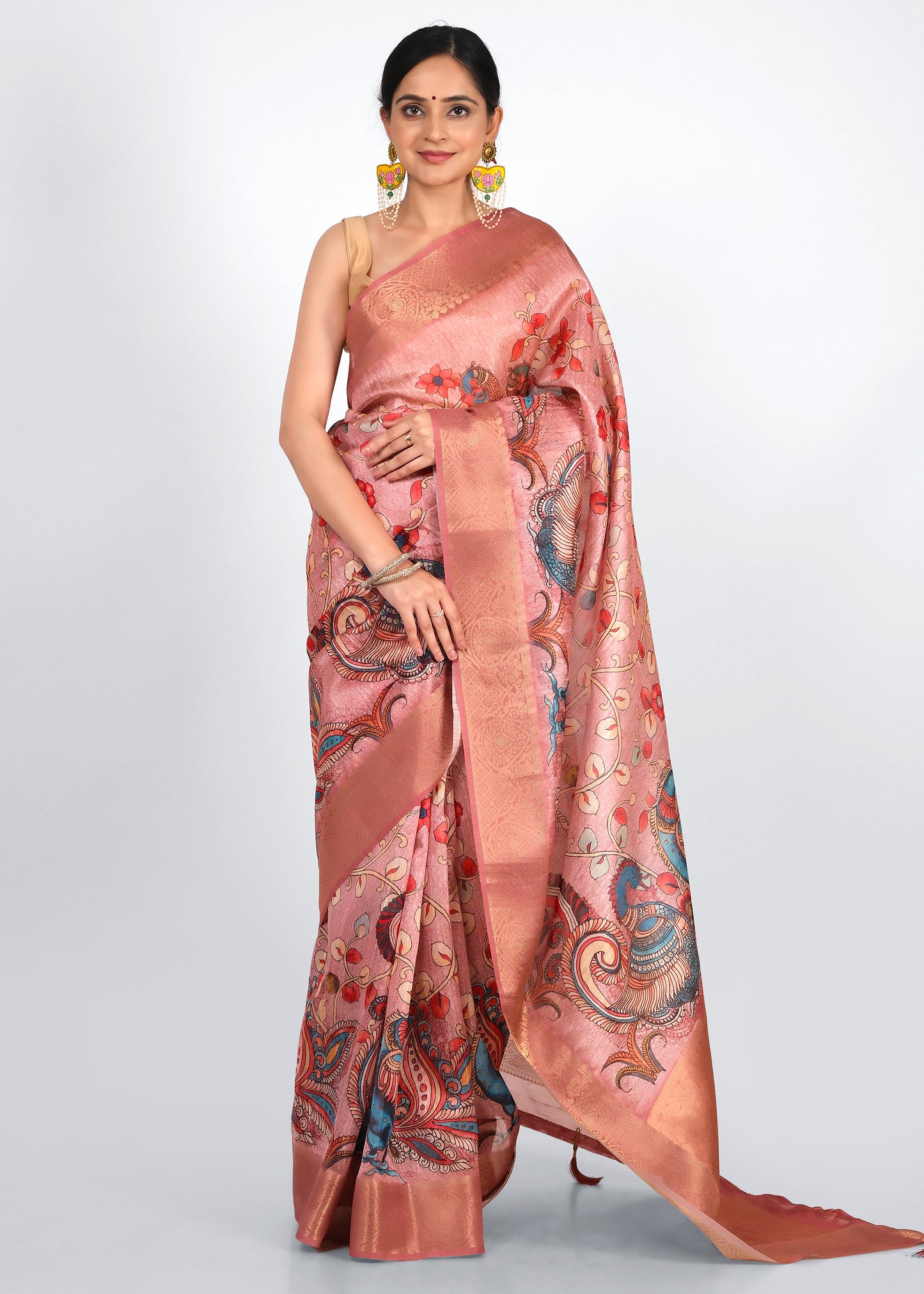 Woman wearing a pink silk saree with intricate peacock and floral motifs, standing elegantly with statement earrings and traditional bangles.