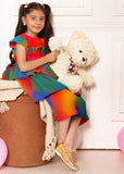 "Girl wearing rainbow pleated dress, holding teddy bear, sitting on a cushioned seat, kids fashion photography."
