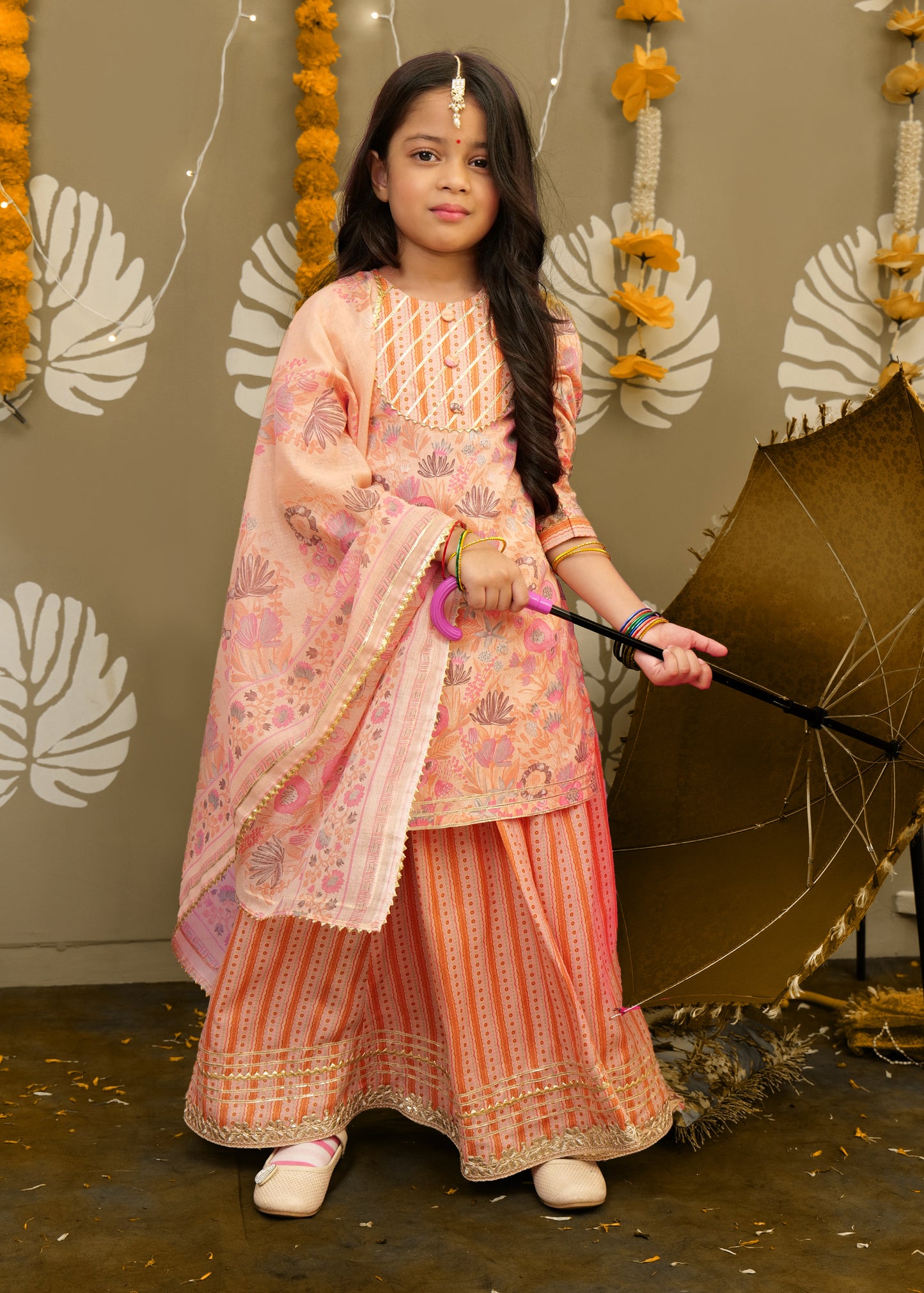A young girl is wearing a peach-colored floral ethnic outfit featuring a flowy skirt and dupatta, accessorized with bangles, and holding a brown traditional umbrella.