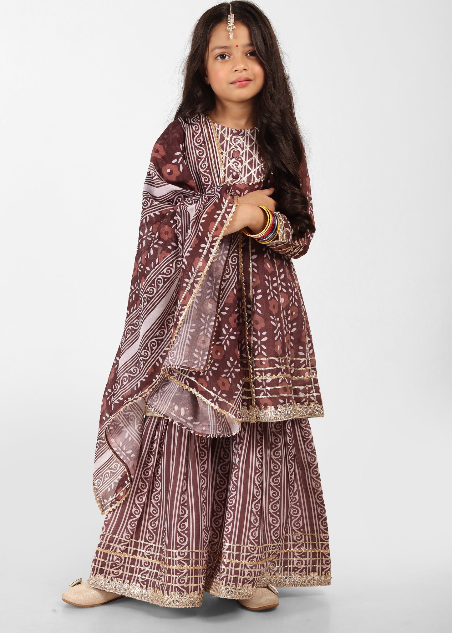"Young girl wearing a stylish maroon ethnic outfit with intricate gold embroidery, posing confidently with a traditional dupatta and matching accessories."
