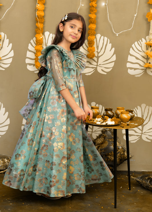 Young girl wearing a teal and gold floral gown with intricate ruffle details, standing beside a table adorned with festive marigold garlands.