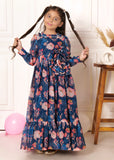 Girl in a blue floral print long dress with puffed sleeves, posing with her hair styled in pigtails, standing near a wooden stool.
