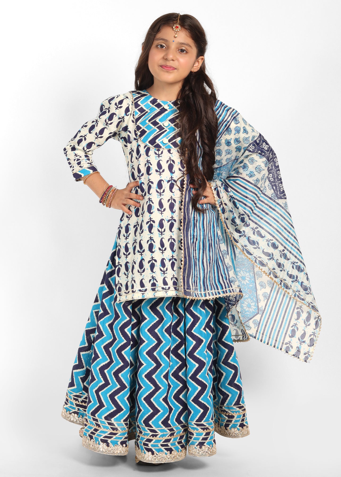 "Girl in a vibrant white and blue printed lehenga choli with zig-zag patterns and floral designs, posing confidently."