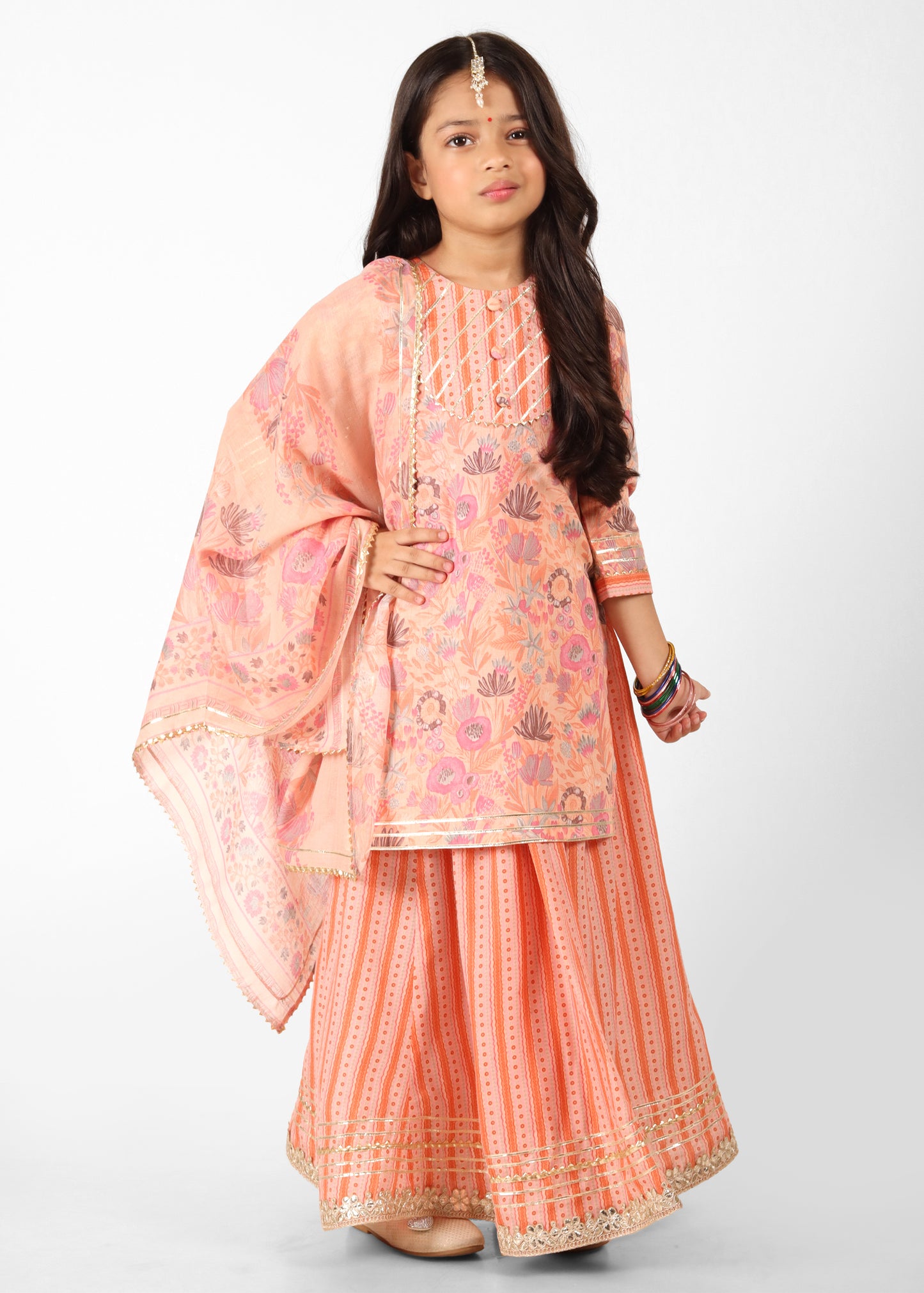 A full-body portrait of a girl wearing a peach-colored lehenga with floral prints, standing against a white background, perfect for festive celebrations.