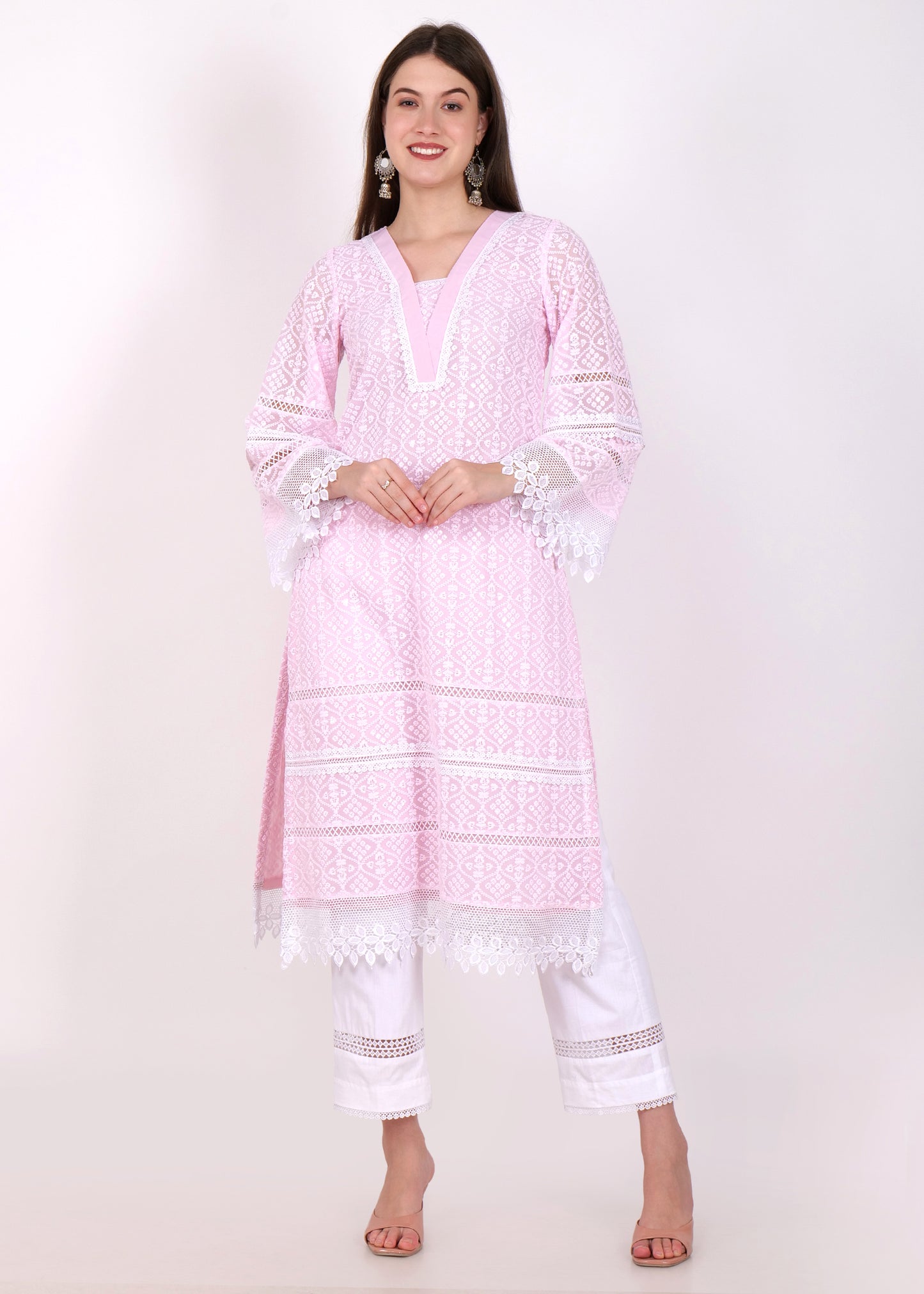 Woman wearing a soft pink embroidered kurta with intricate white lace detailing, styled with white pants, posing with hands clasped