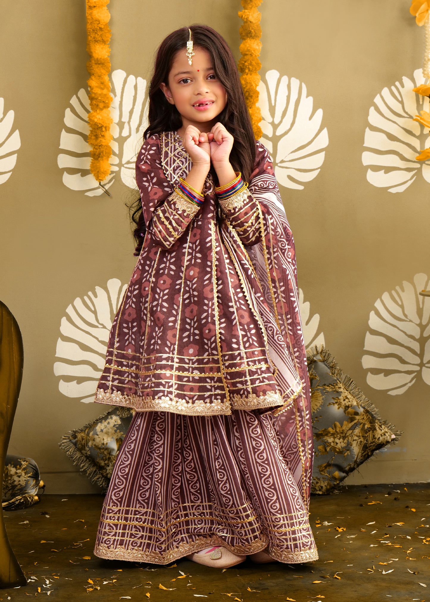A young girl dressed in a maroon ethnic kurta and matching palazzo set with gold detailing, standing in a festive environment. The outfit's floral and geometric prints make it ideal for traditional occasions.