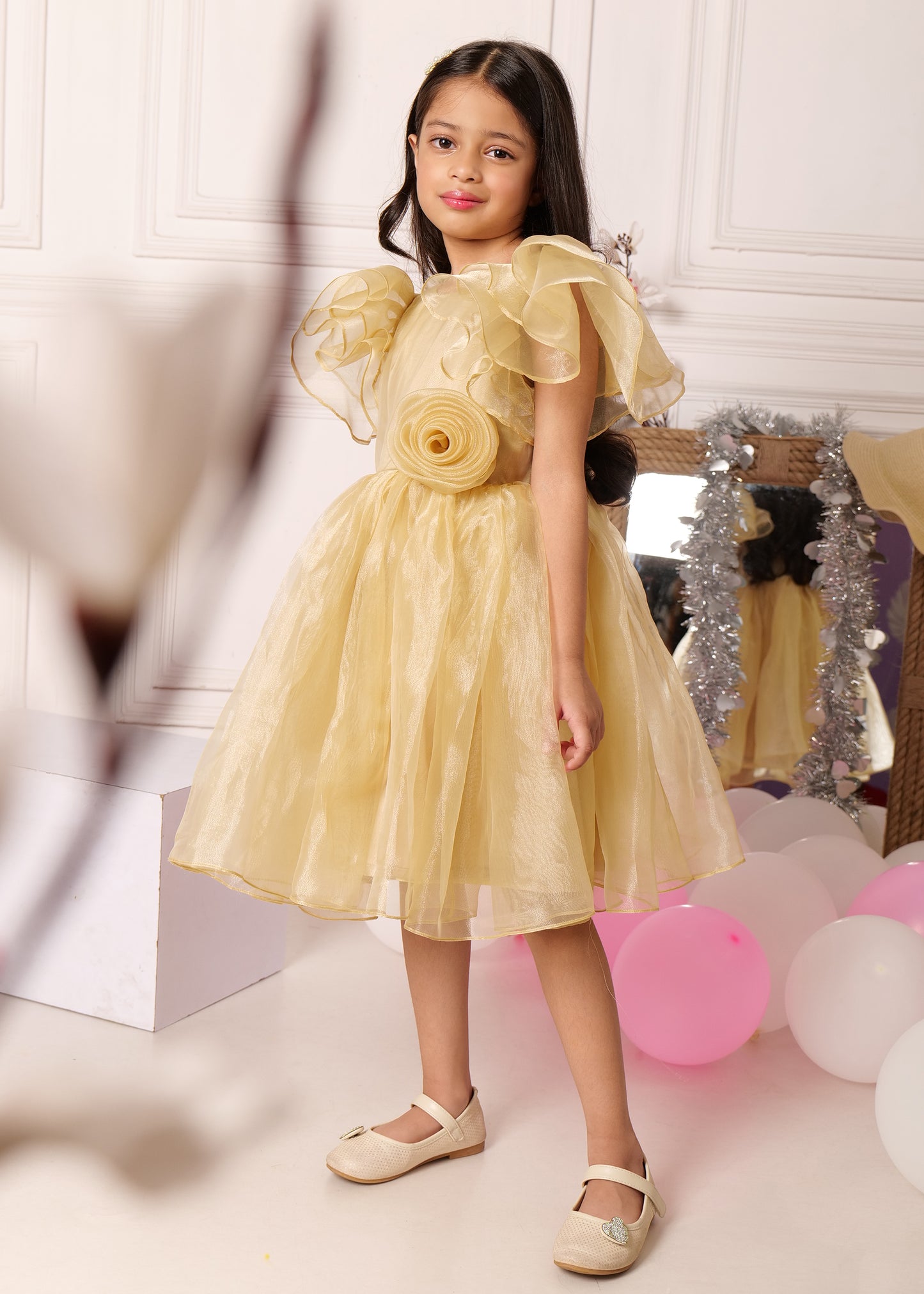 "Side view of a girl in a gold organza dress with layered puff sleeves, standing gracefully by the balloons and reflecting in a mirror."