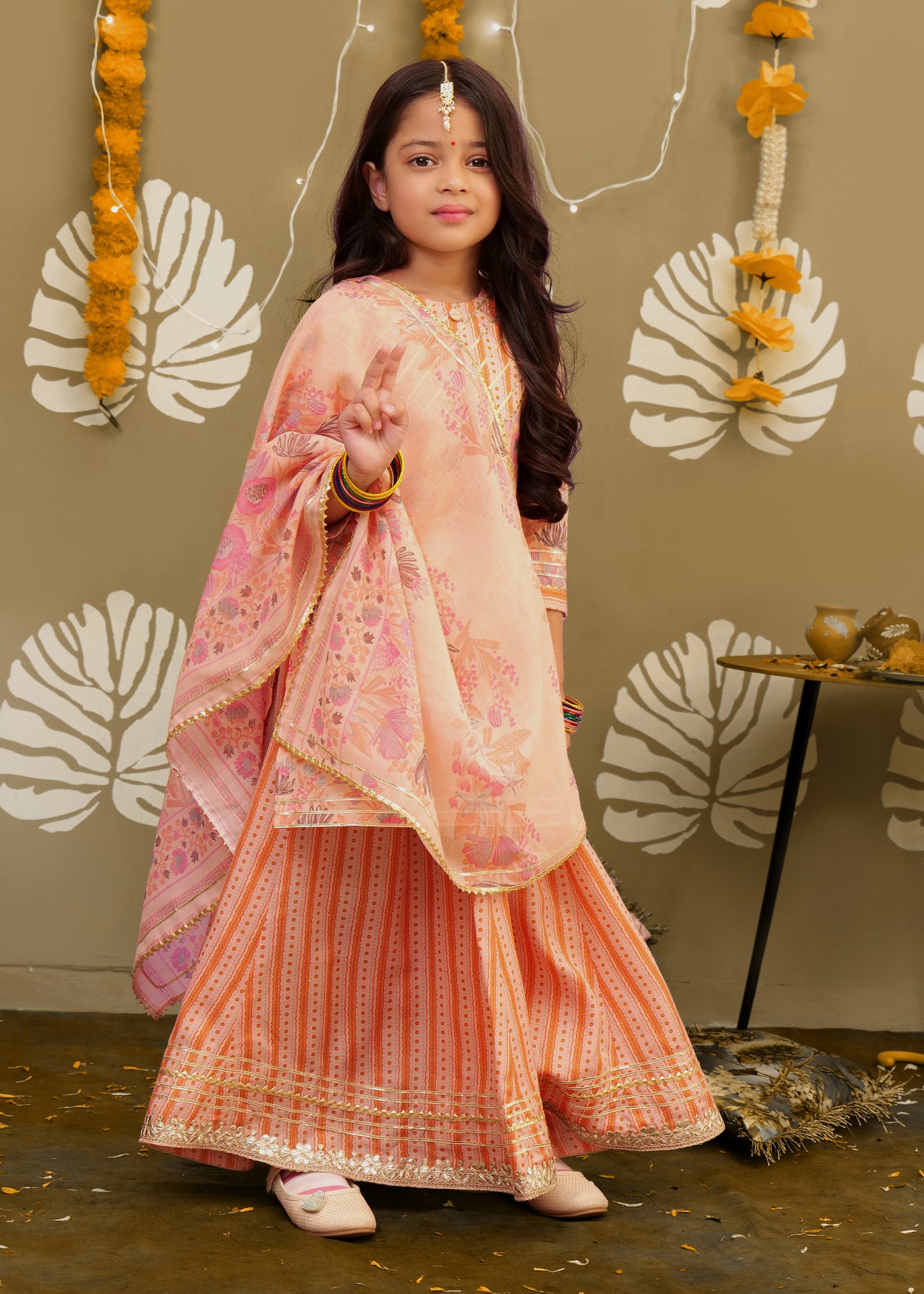 A girl dressed in a peach ethnic lehenga set with floral patterns and a printed dupatta, adorned with bangles and traditional accessories, posing against a festive backdrop.