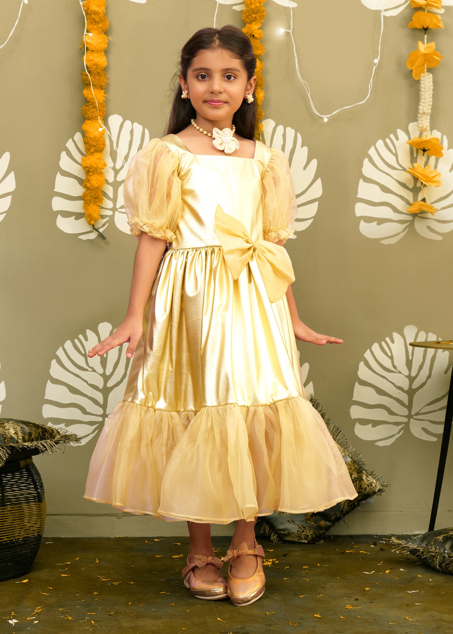 A child model wearing a gold puff-sleeve dress with a large waist bow, posing gracefully in front of festive floral décor.