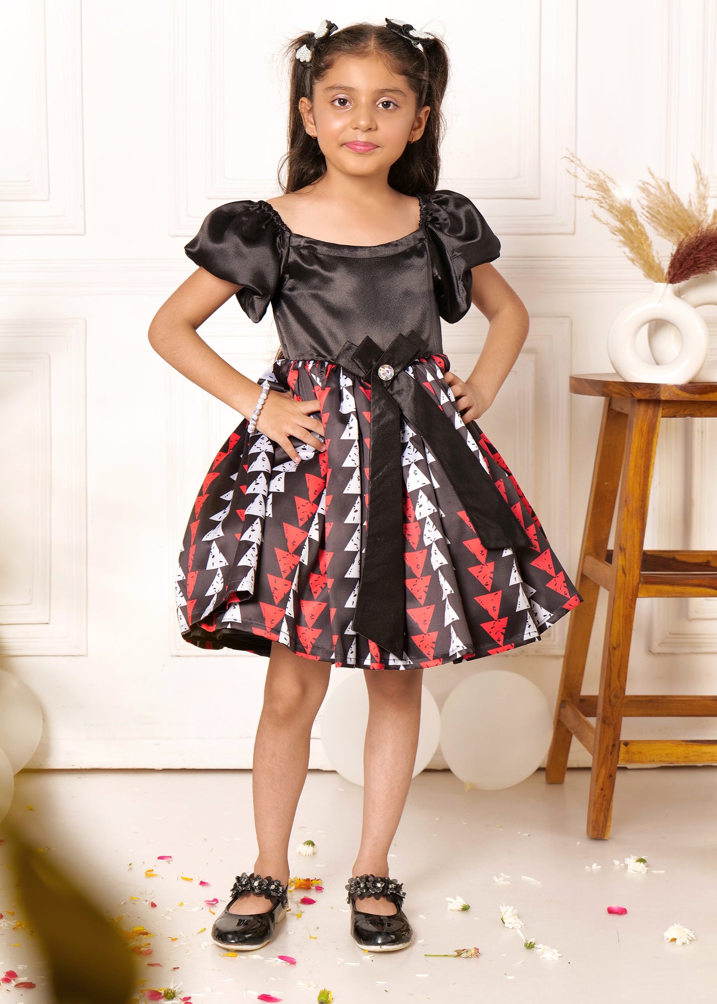 Young girl posing in a black satin dress with red and white triangle prints and a large bow at the waist, showcasing kids' fashion trends