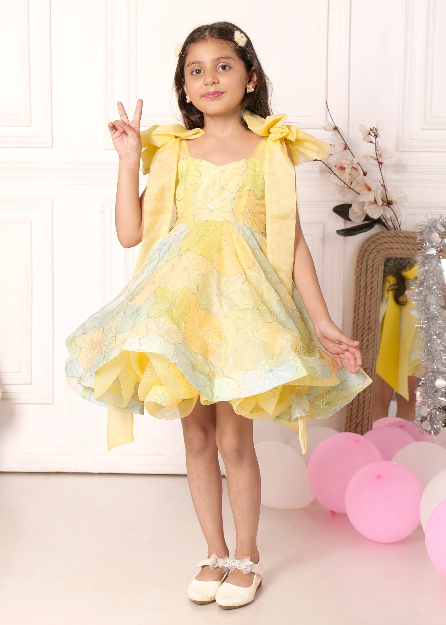 "Smiling girl in a pastel yellow floral dress with bow detailing on the straps, showing off her playful side while making peace signs."