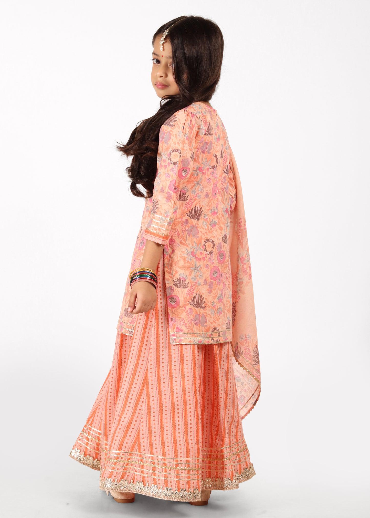 Back view of a girl dressed in a peach lehenga set with floral prints, showing the intricate detailing of the fabric and flowy silhouette.