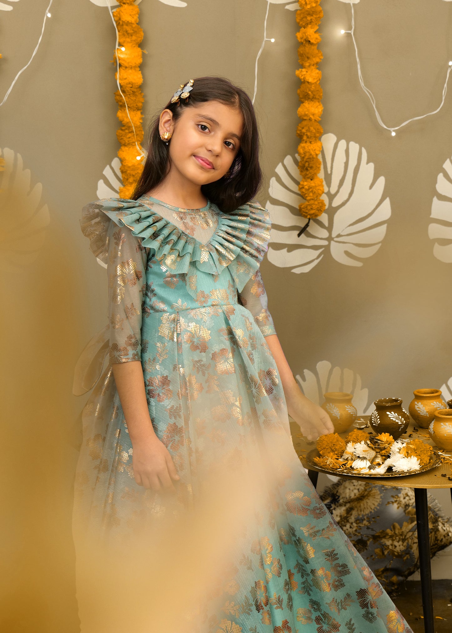 A little girl in a teal and gold floral gown with statement ruffle details, standing in a festive setting decorated with marigolds.