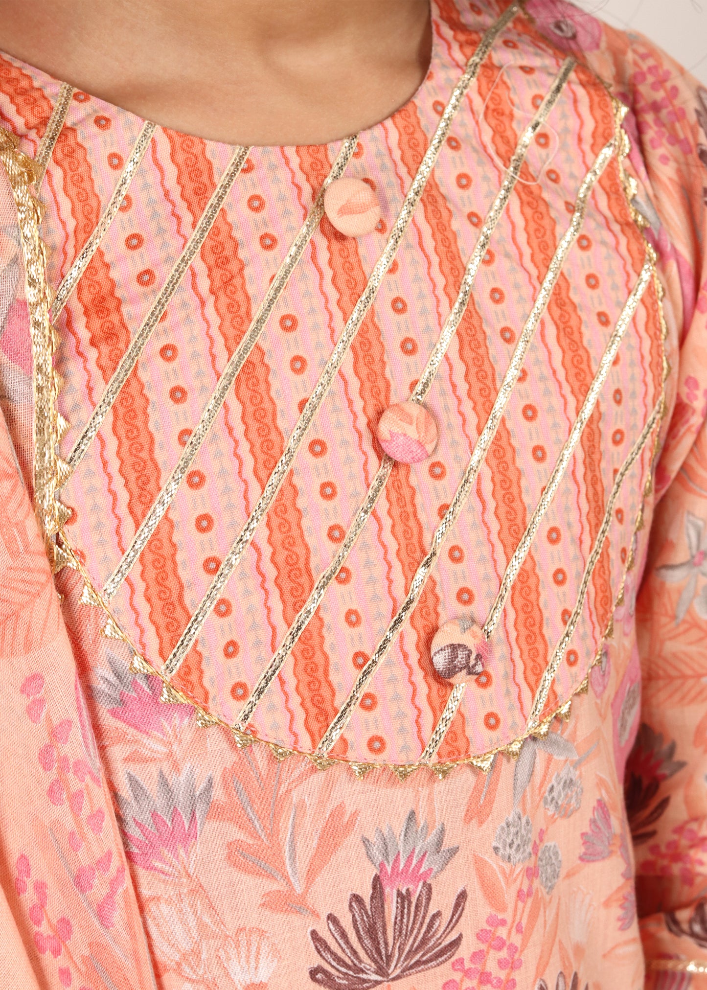 A detailed close-up of the intricate embroidery on the bodice of a peach-colored lehenga set designed for young girls, with gold highlights and floral motifs.