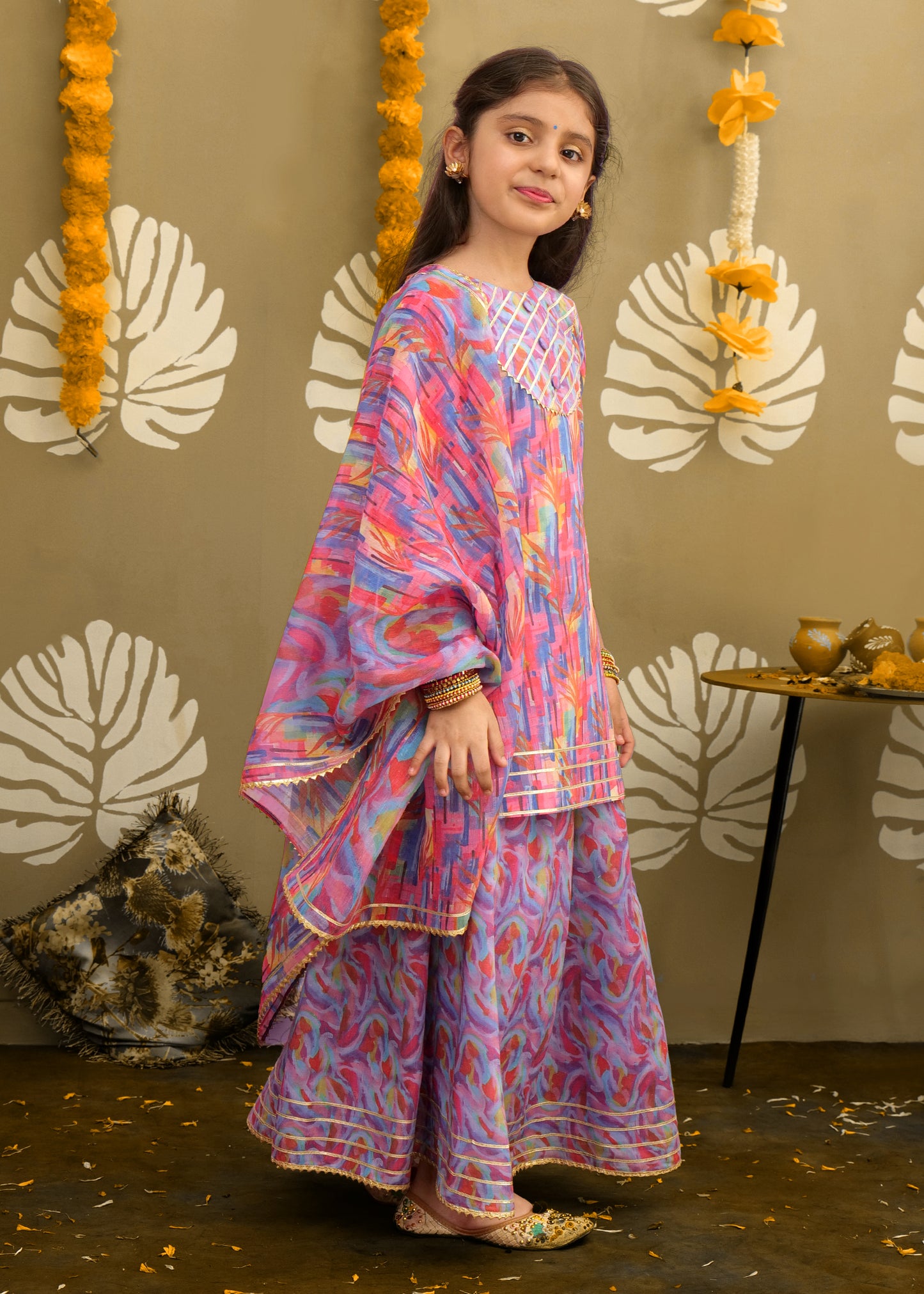 The girl posing elegantly in a bright, multi-colored traditional dress, accessorized with gold earrings and bangles, ideal for festive and cultural events.