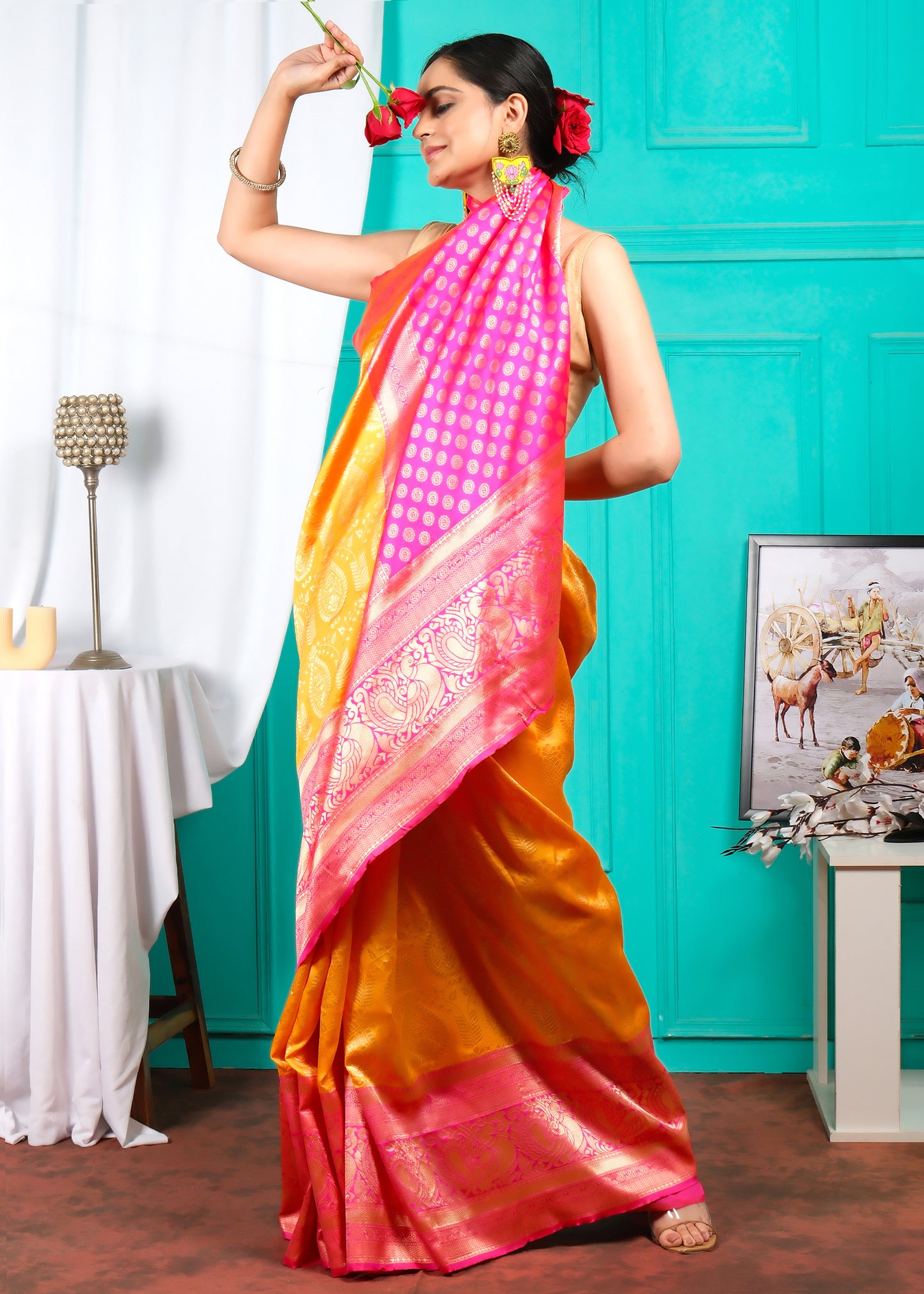 Woman in a festive orange silk saree with a pink pallu, holding roses, styled with colorful earrings and a playful pose