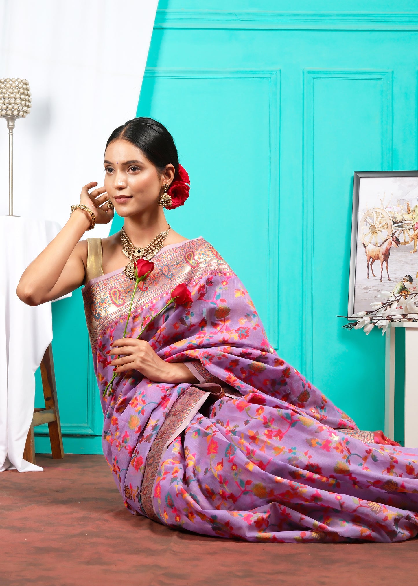 Woman seated in a lavender floral saree, holding a red rose, elegantly adorned with traditional gold jewelry and floral hair accents against a teal backdrop