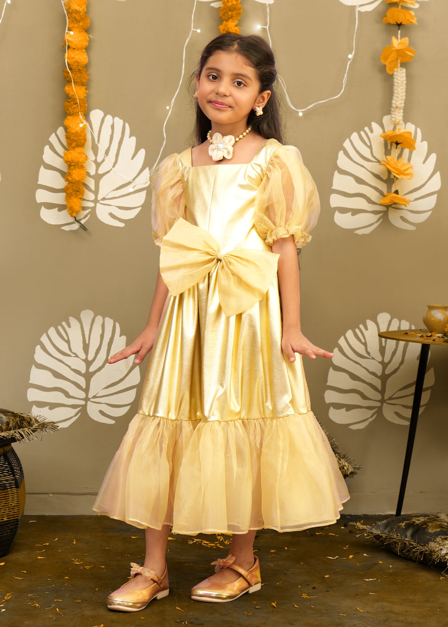 A girl in a golden gown with puffed sleeves and a ruffled hemline, posing confidently in front of an artistic backdrop with festive garlands.