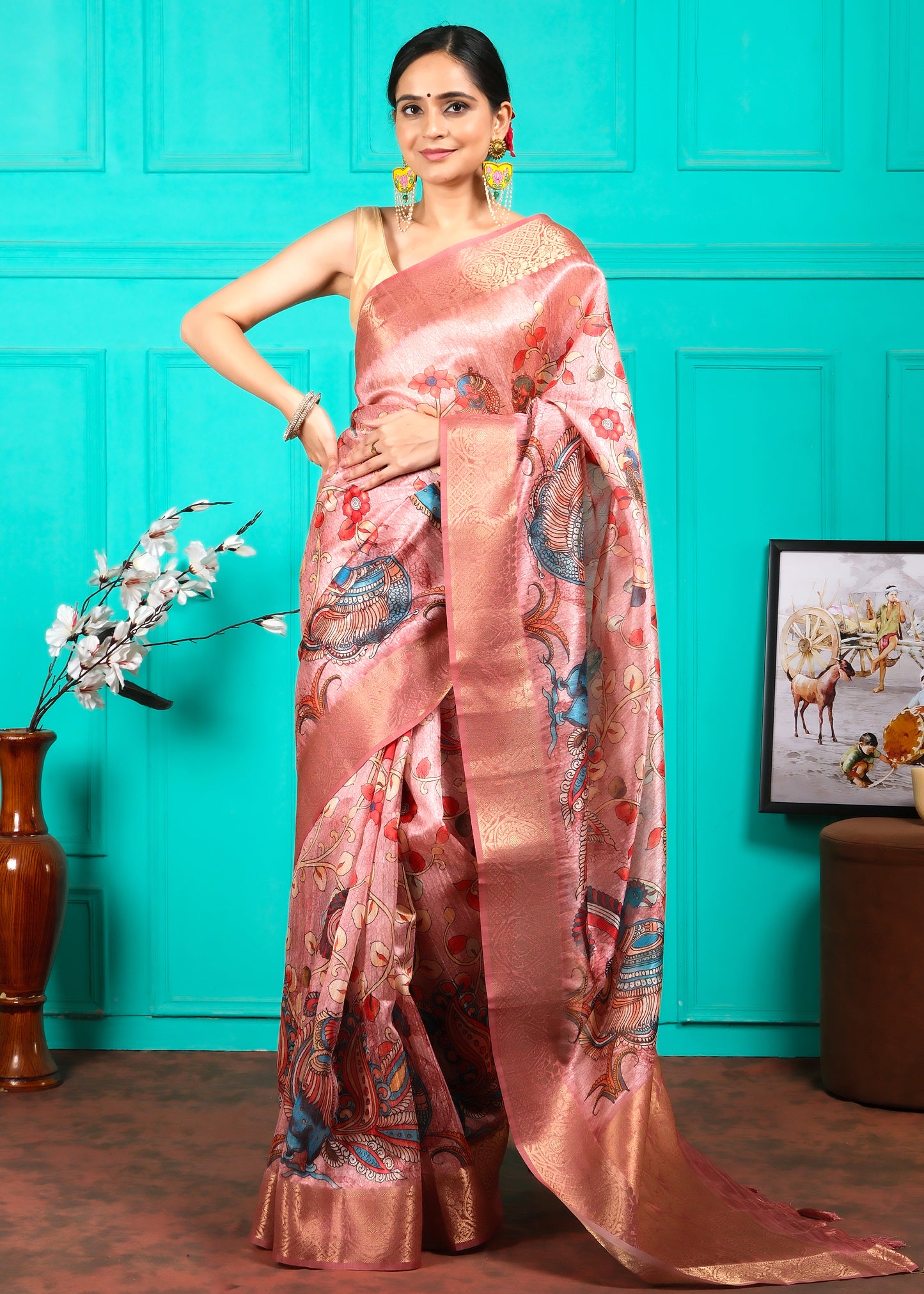 Model in a traditional pink silk saree with detailed floral and peacock patterns, posing against a turquoise backdrop for a festive look.