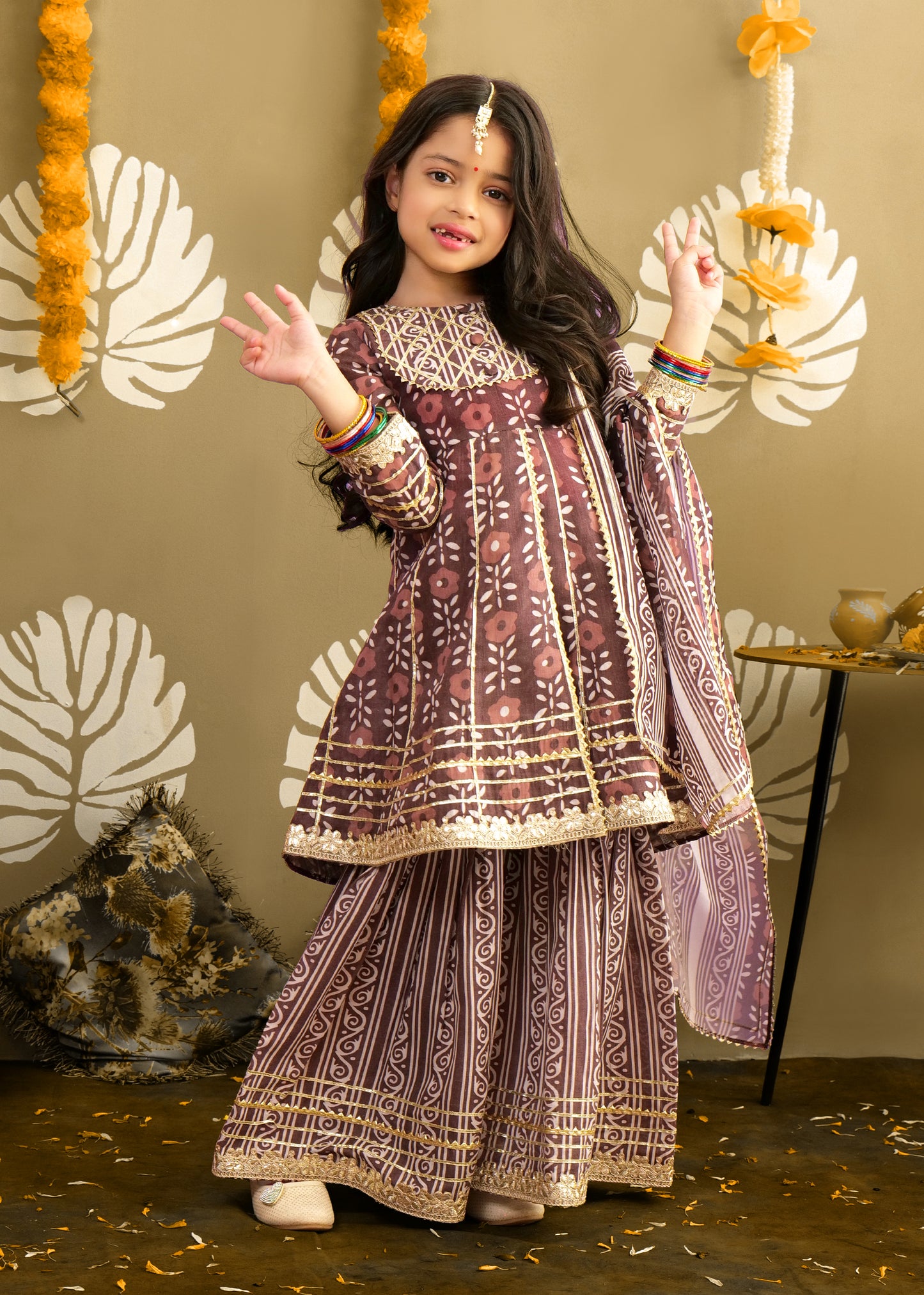A young girl wearing a traditional maroon printed kurta and palazzo set, posing joyfully in a festive setting. The outfit features intricate gold embroidery and floral patterns, perfect for Indian celebrations.