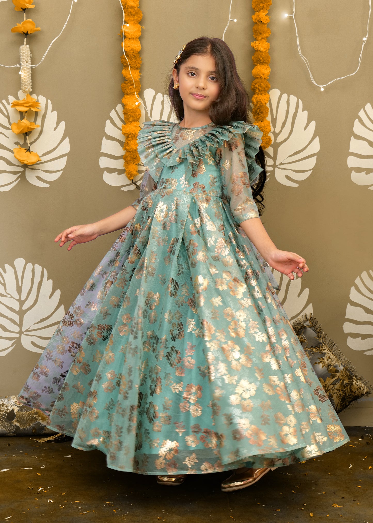 Young girl posing in a teal floral gown with golden accents, holding the dress’s full skirt in a joyful twirl.