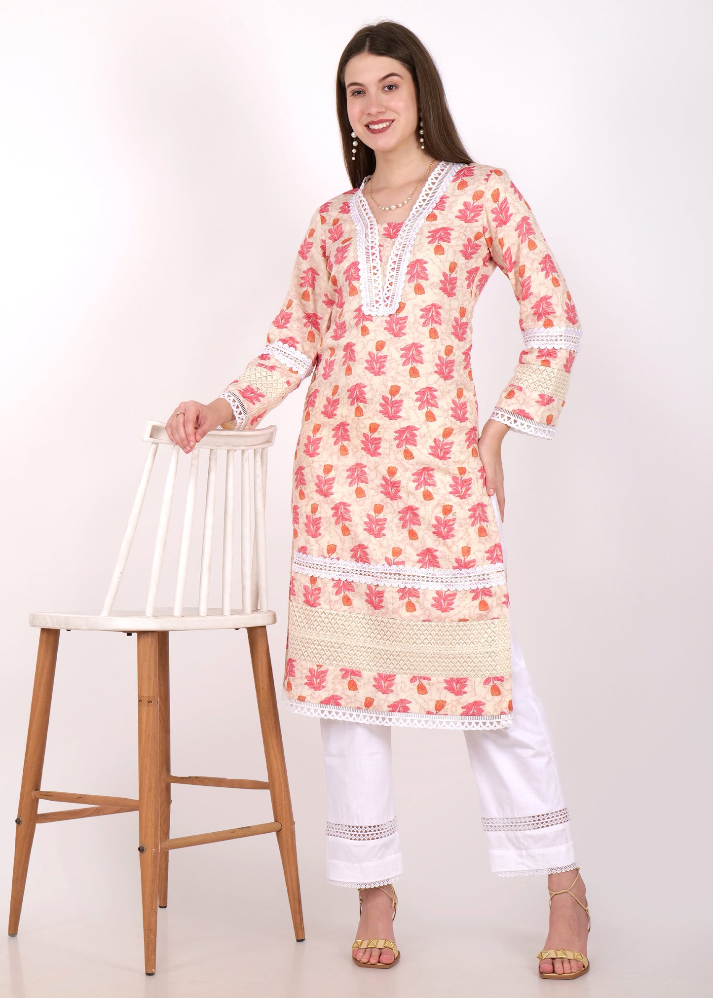 Woman wearing a beige and pink floral printed kurta with white lace detailing, standing near a chair, paired with white pants