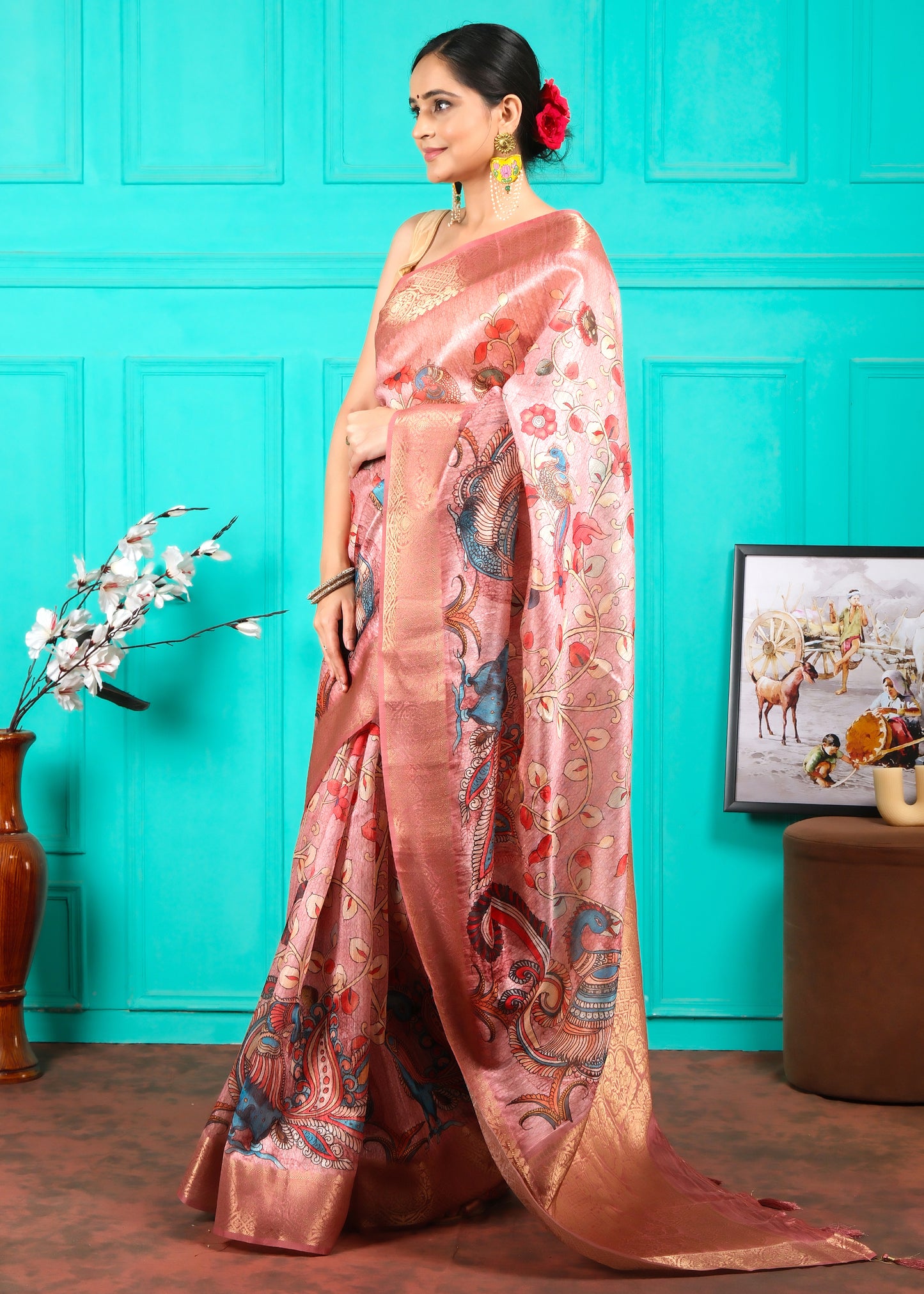 Woman adorned in a pink silk saree with delicate gold work and colorful motifs, posing with flowers in her hair and radiant accessories
