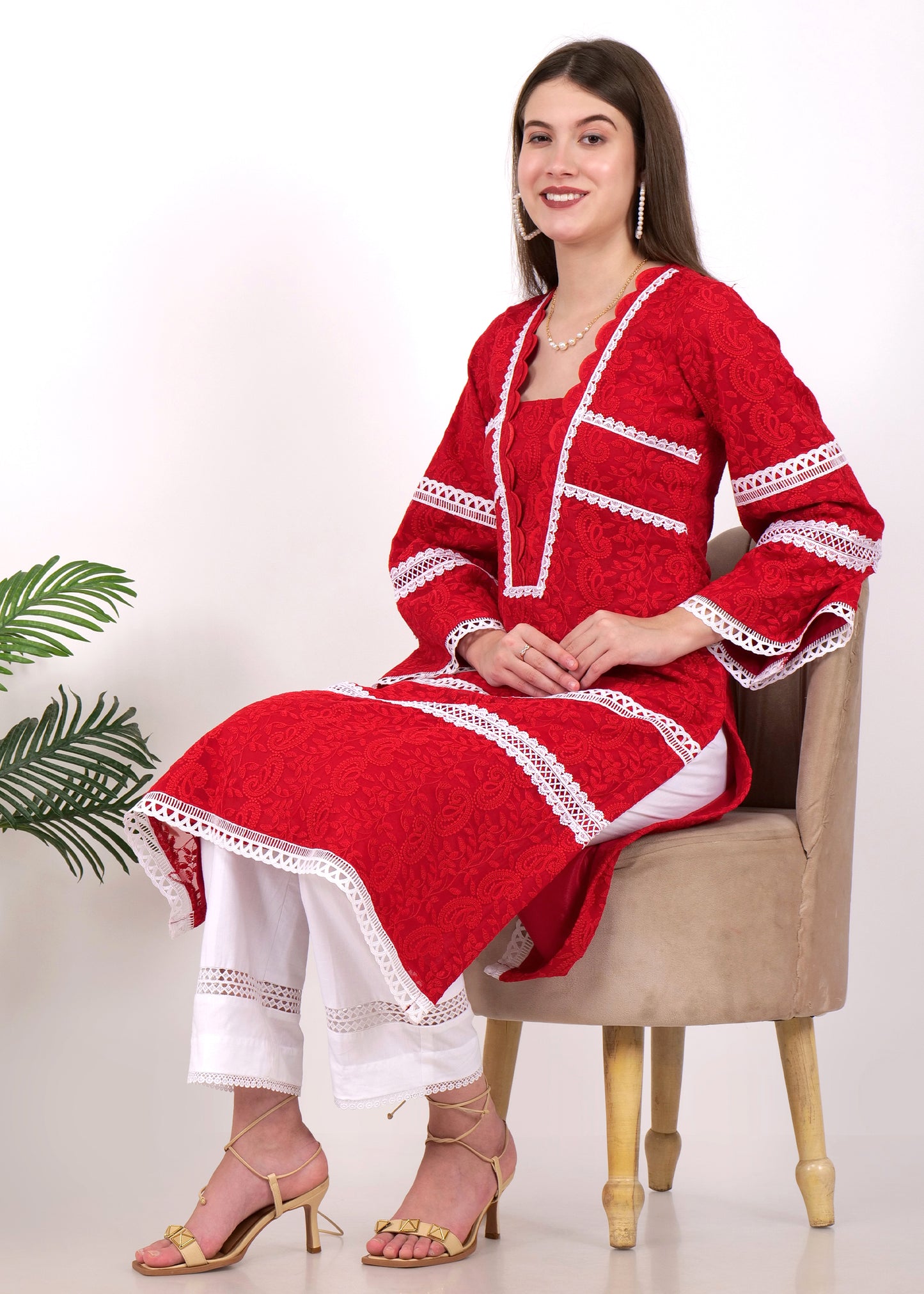 The model sitting gracefully in a red lace kurta set with white lace accents, paired with white pants and gold heels