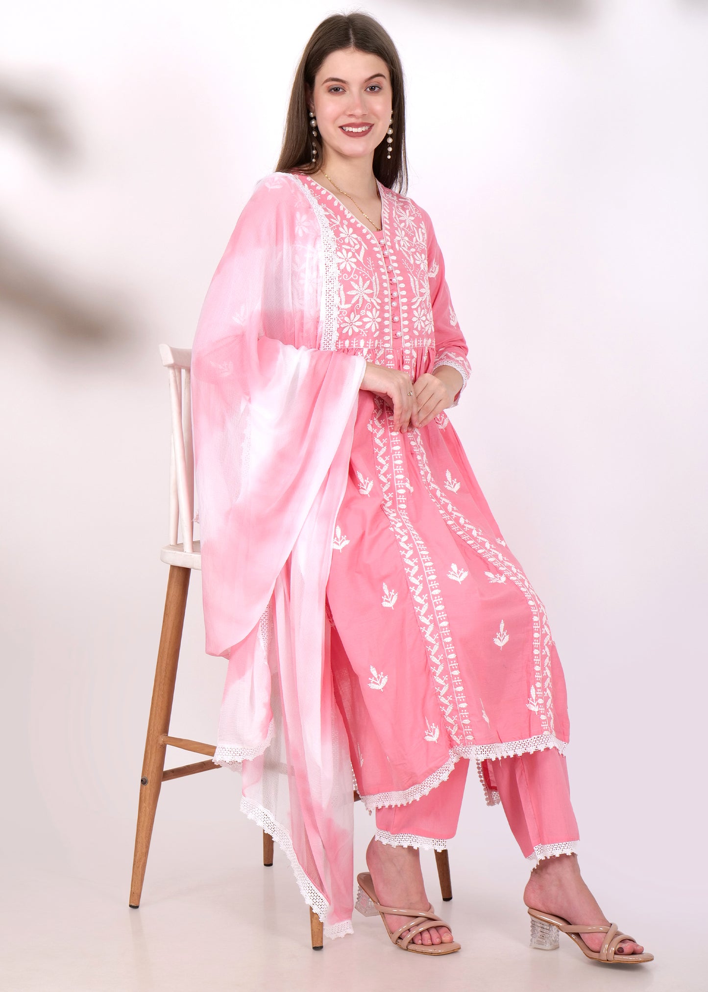 Woman sitting gracefully, wearing a pink embroidered Anarkali suit with white floral detailing and a matching sheer dupatta.