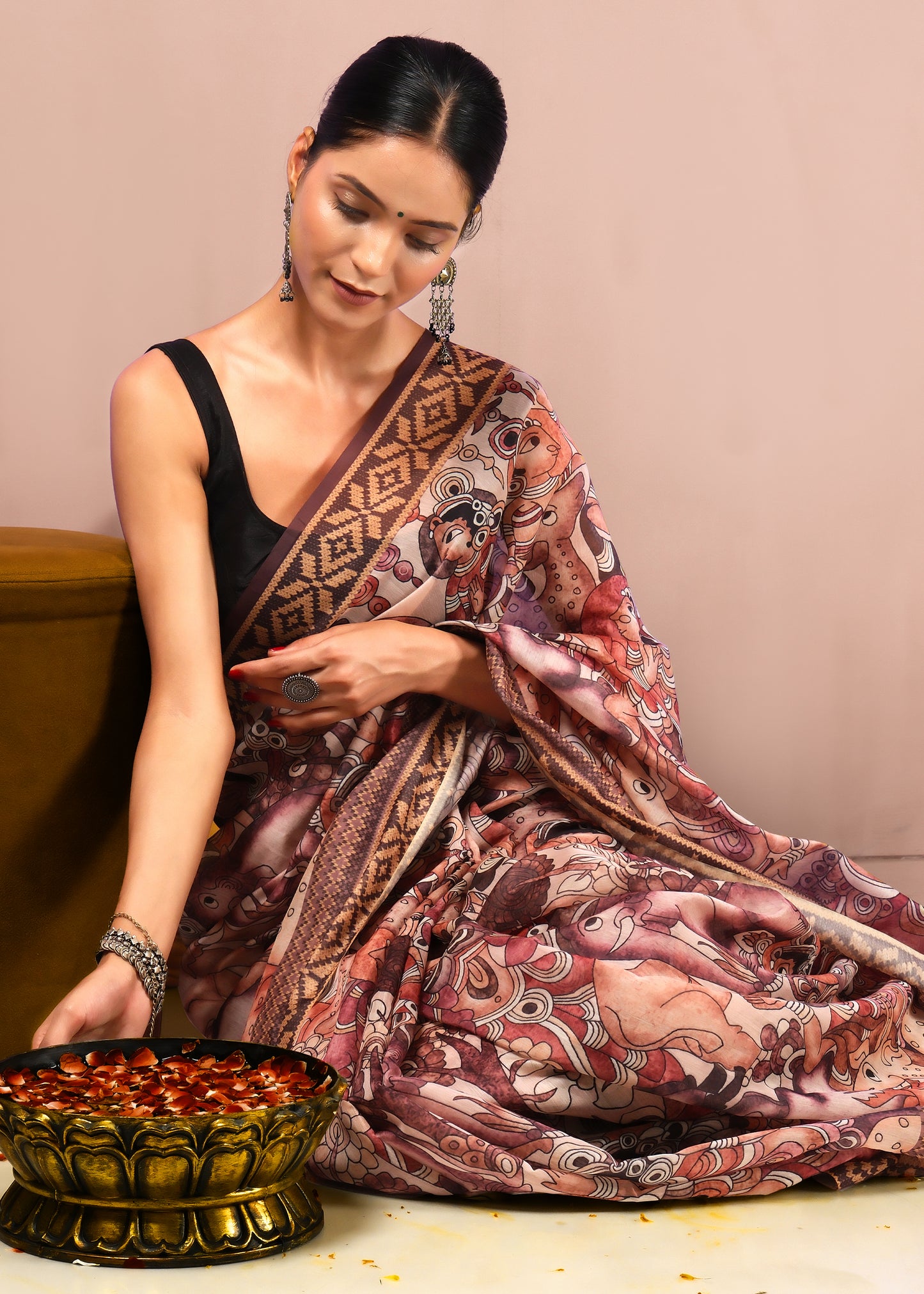 "A woman seated gracefully in a saree with mythological art, engaging in a ritual, set in a traditional decor environment."