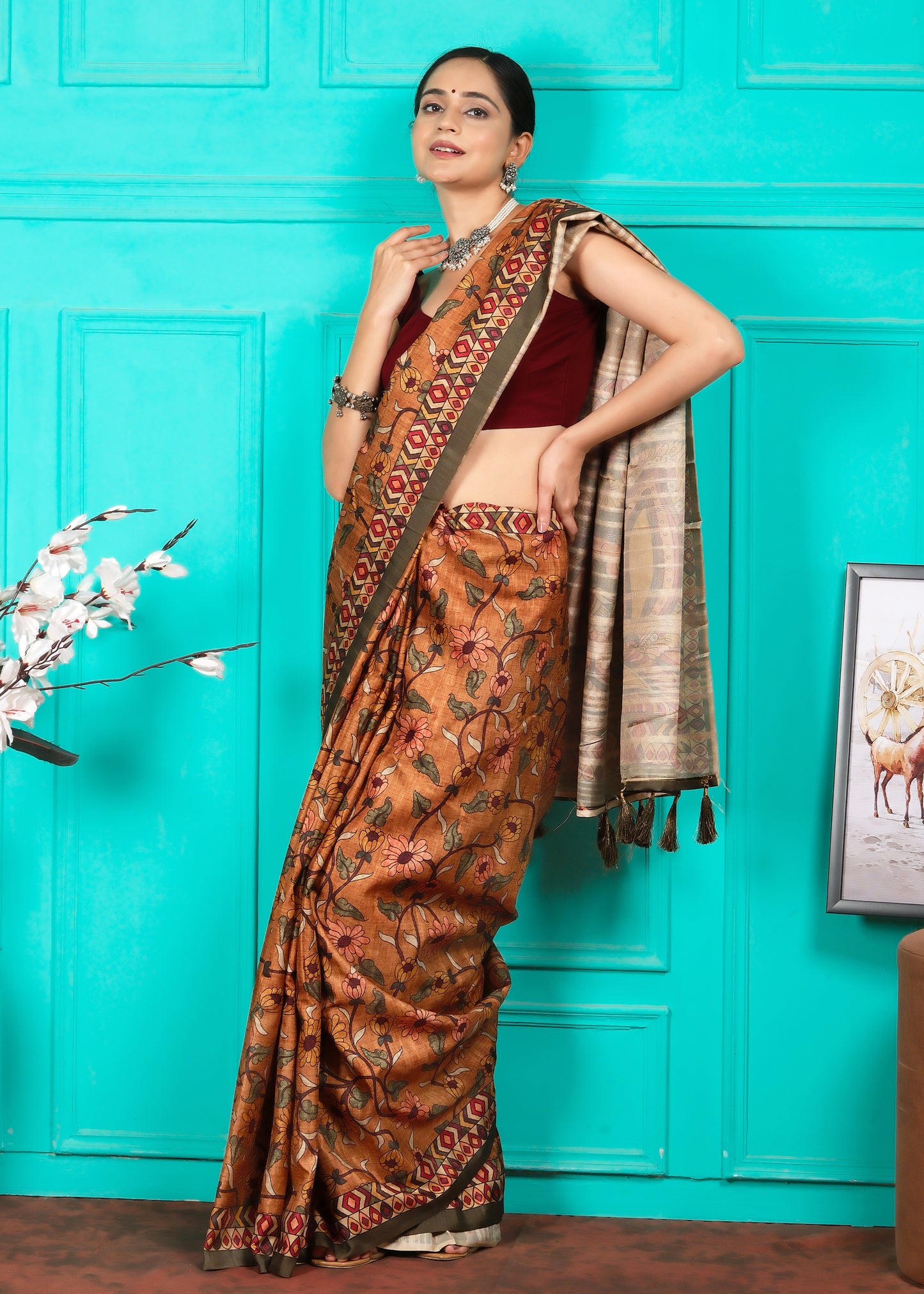 A front-facing view of a woman dressed in a floral-patterned handloom saree with a vibrant geometric border, standing in a tastefully decorated room. The bright turquoise background and her ethnic jewelry accentuate the richness of the saree's design.
