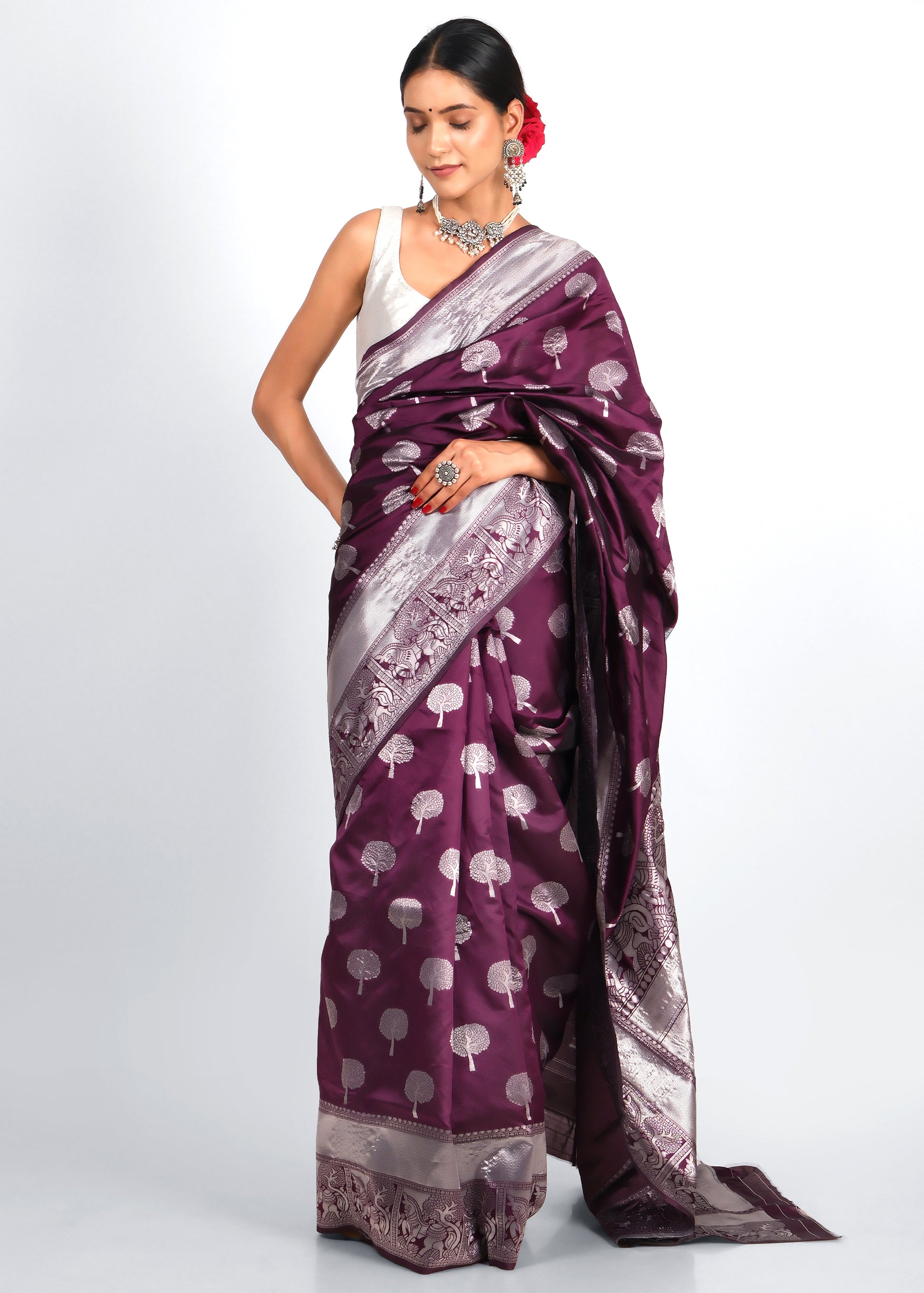 Model in Purple and Silver Banarasi Silk Saree with Tree Motif Embroidery
The model is gracefully showcasing a traditional purple and silver Banarasi silk saree, adorned with intricate tree motifs and broad silver borders, paired with silver jewelry and a red floral hair accessory.