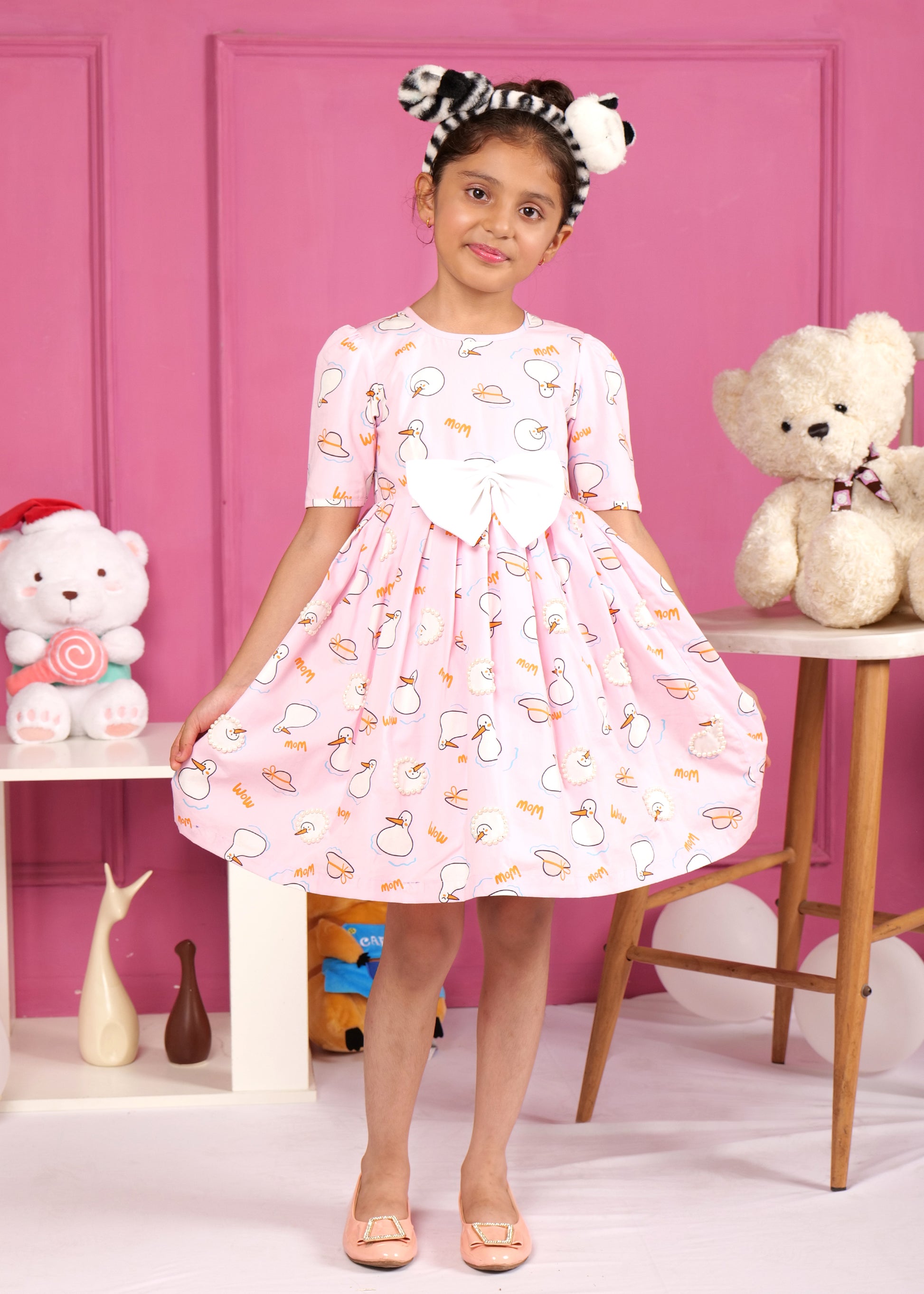 Little girl wearing a pink dress with swan print, accessorized with a cute bow headband, posing in front of a pink wall surrounded by toys