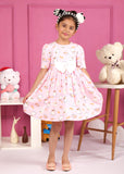 Little girl wearing a pink dress with swan print, accessorized with a cute bow headband, posing in front of a pink wall surrounded by toys