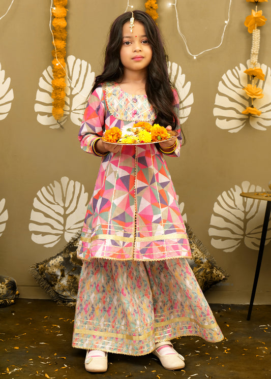 A young girl dressed in a colorful geometric patterned kurta with a floral palazzo, holding a marigold flower thali. The outfit features bright patterns, making it perfect for Indian festive occasions.