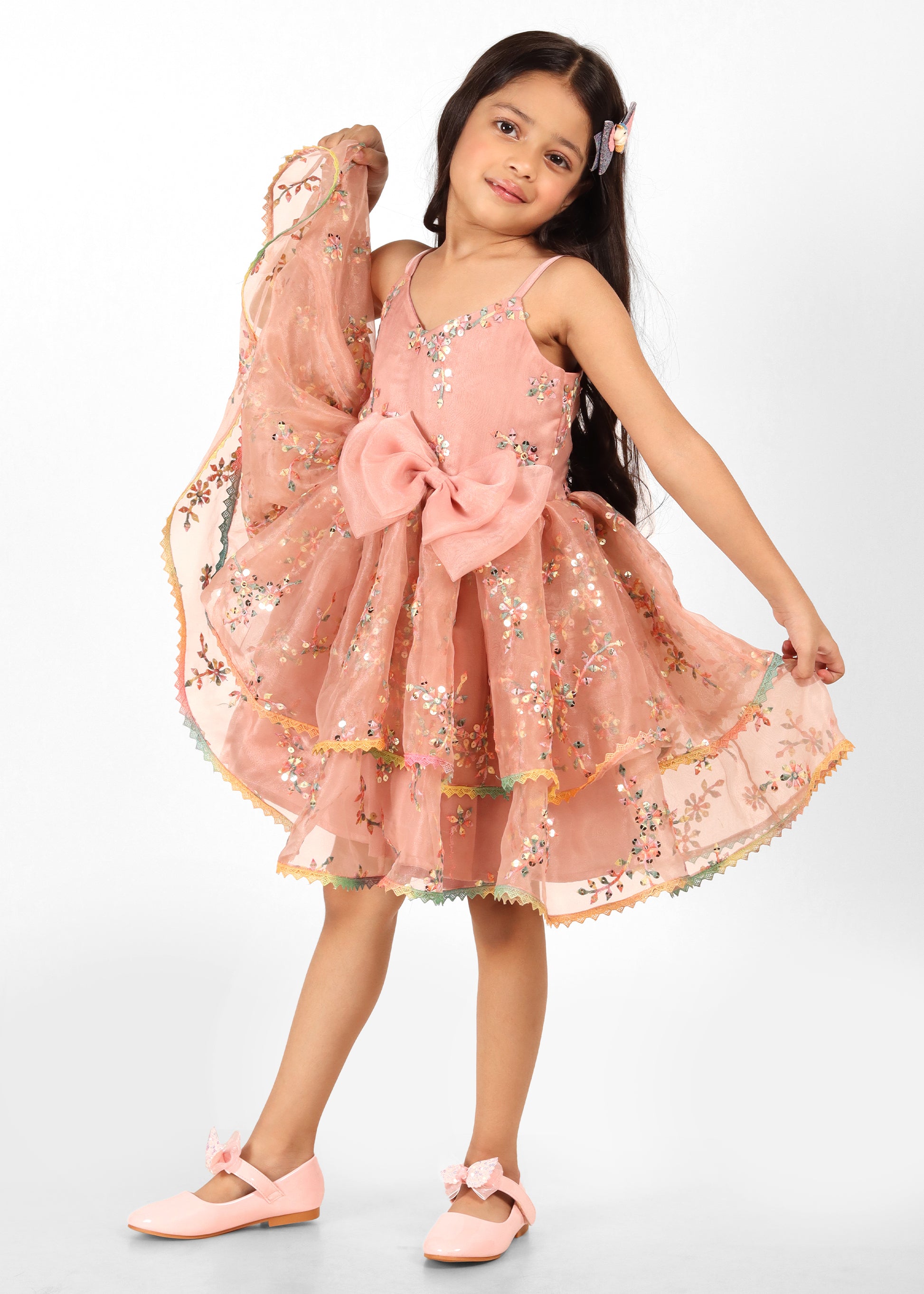 A girl showcasing a peach floral dress with layers of tulle, holding the dress elegantly to highlight its intricate details and feminine design.