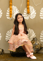 A young girl wearing a peach floral dress with sequin detailing and a big bow, posing elegantly while sitting on a basket with decorative cushions in the background.