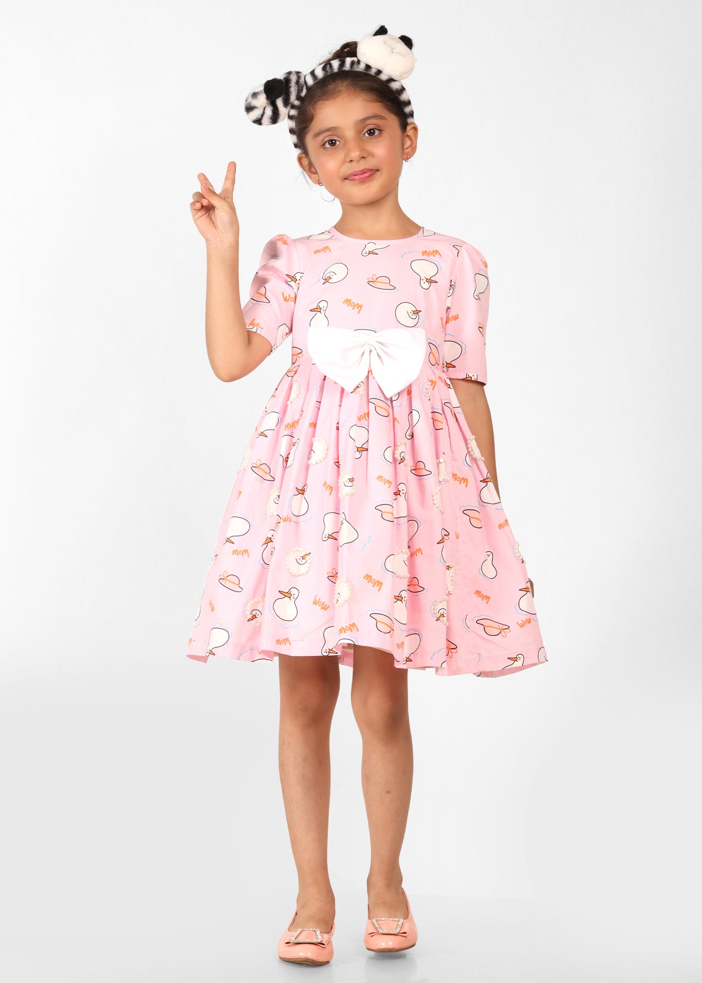 Girl wearing a light pink dress with white bows and cute animal prints, striking a peace sign with her hand, perfect for casual and festive occasions