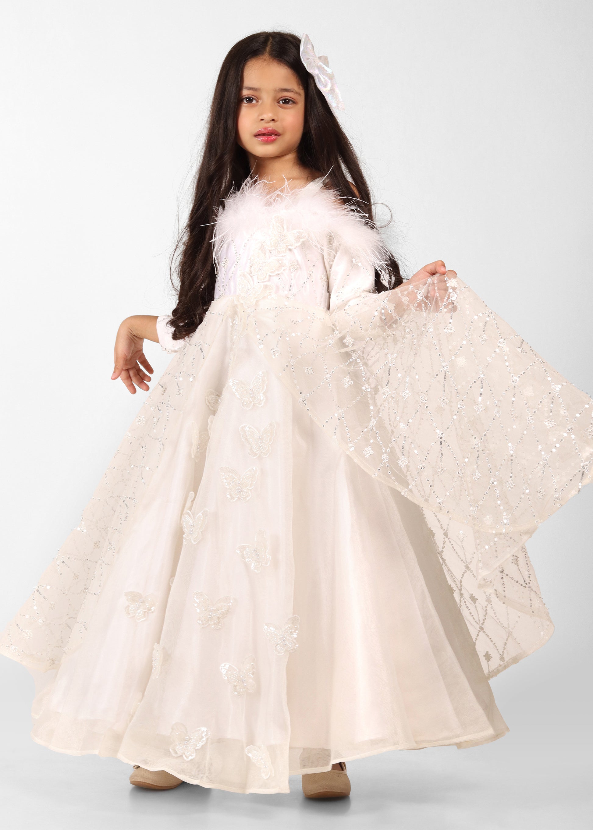 Little girl wearing a white butterfly-sequined gown with long sleeves and feather detailing, standing elegantly in a white room.