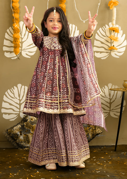  A young girl wearing a traditional maroon printed kurta and palazzo set, posing joyfully in a festive setting. The outfit features intricate gold embroidery and floral patterns, perfect for Indian celebrations.