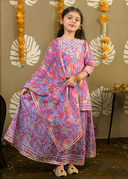 A young girl dressed in a vibrant multi-colored traditional outfit, with pink, blue, and gold detailing, showing a confident pose at a festive event.