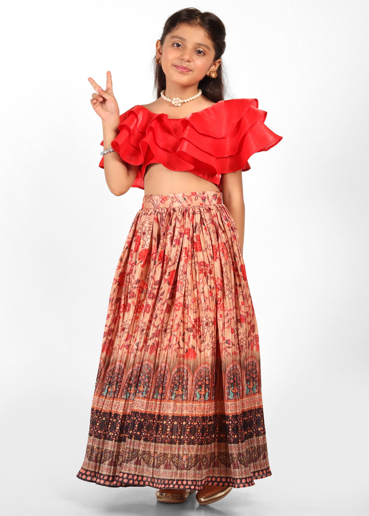 A young girl wearing a vibrant red ruffle top paired with a printed long skirt featuring floral and traditional motifs. She poses confidently with a peace sign.