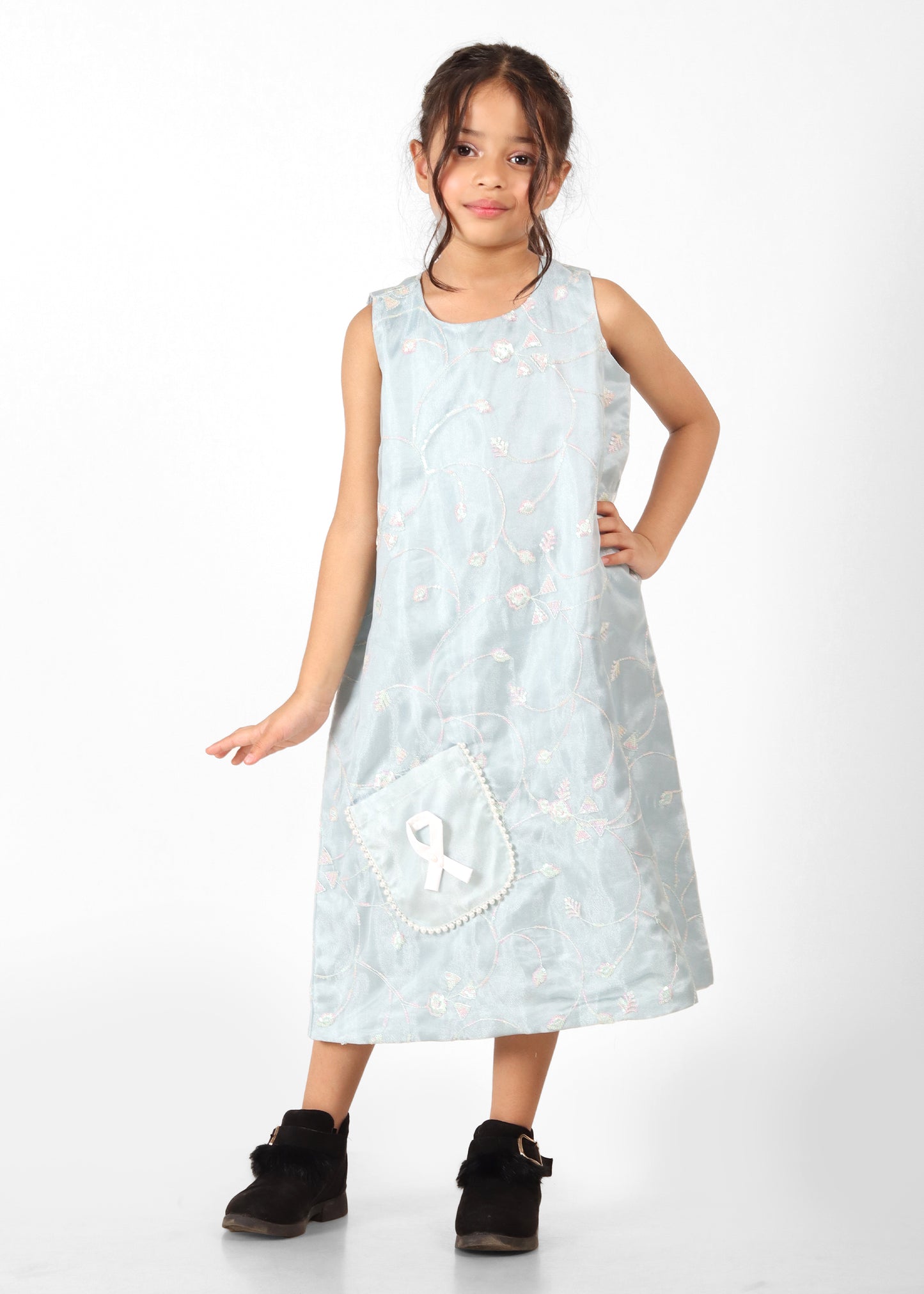 Girl wearing a light blue embroidered sleeveless dress with a white ribbon detail on the pocket, posing confidently next to a wooden stool