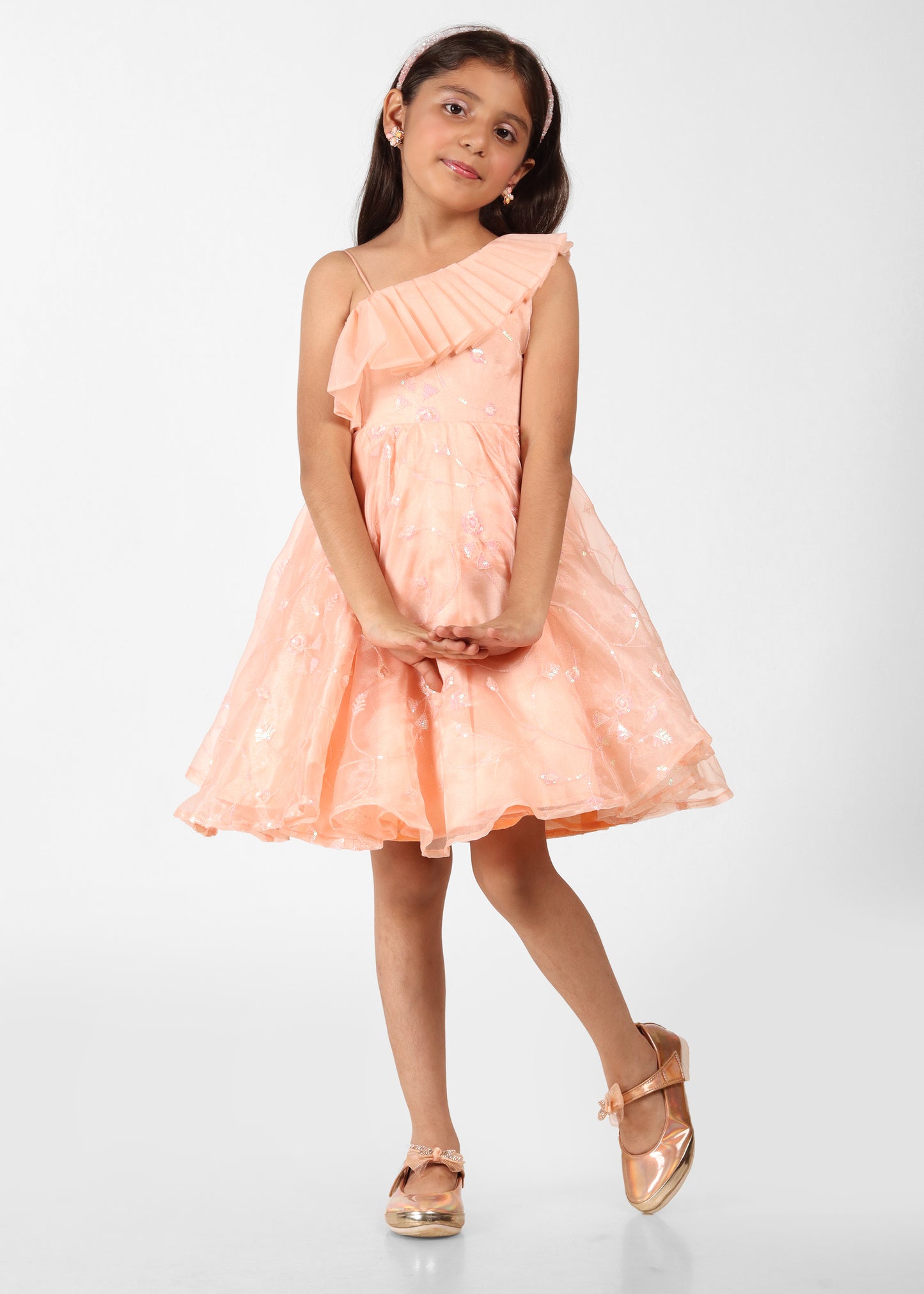 "A young girl wearing a peach one-shoulder dress with a pleated ruffle detail and subtle floral patterns, posing with a gentle smile and her hands clasped in front."