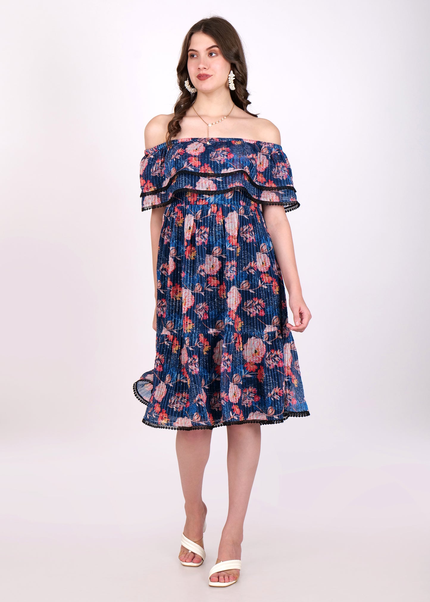 Woman wearing an off-shoulder navy blue floral pleated midi dress with ruffled layers and black lace trim detailing, styled with white sandals.